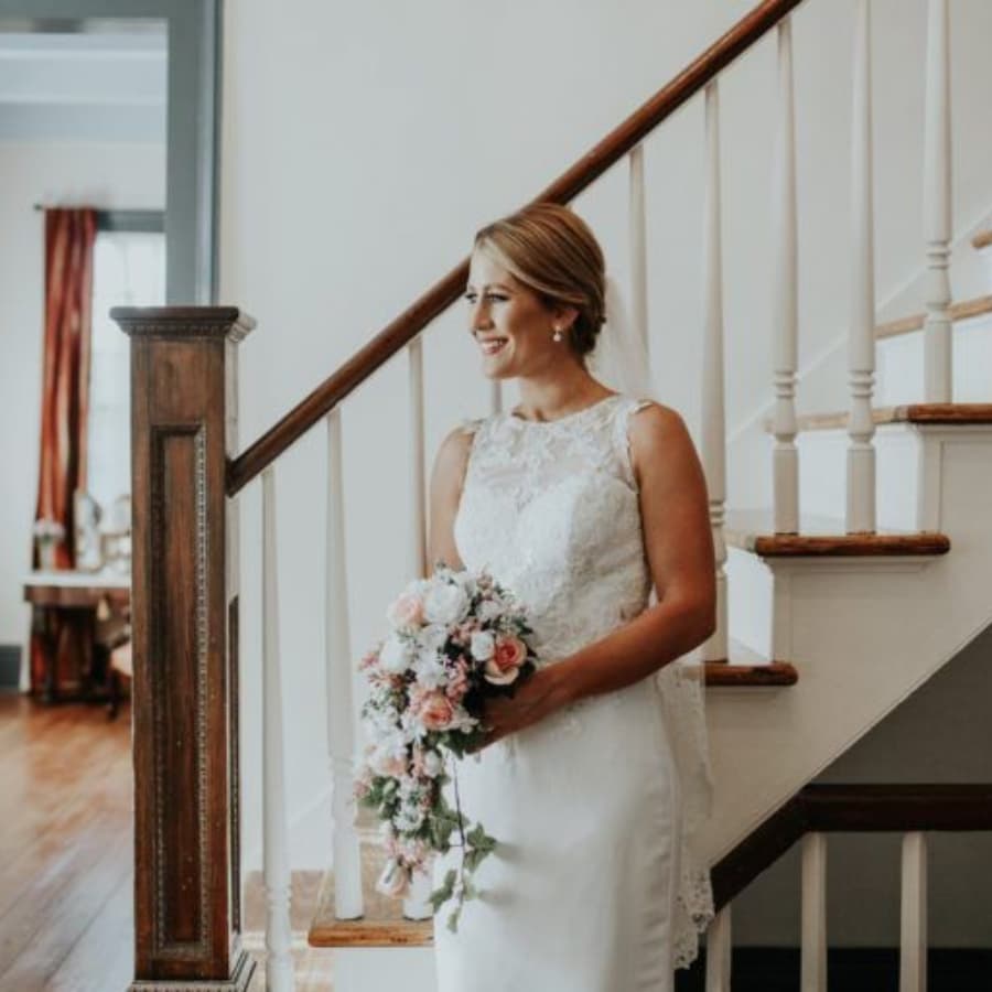 A bride in a lace gown smiles while holding a floral bouquet near a staircase.