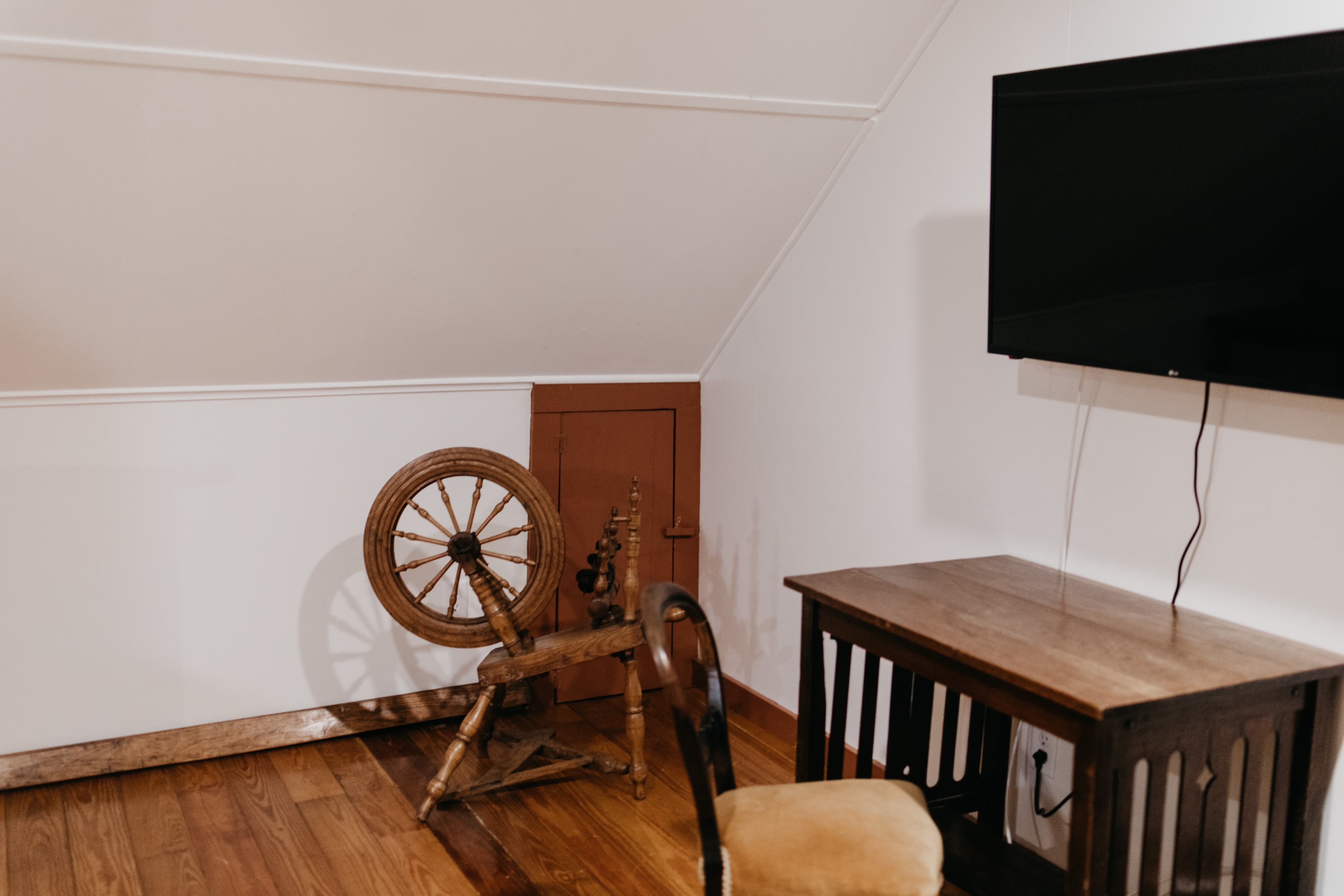 A cozy attic corner featuring a spinning wheel, a wooden desk, and a mounted TV on the wall.