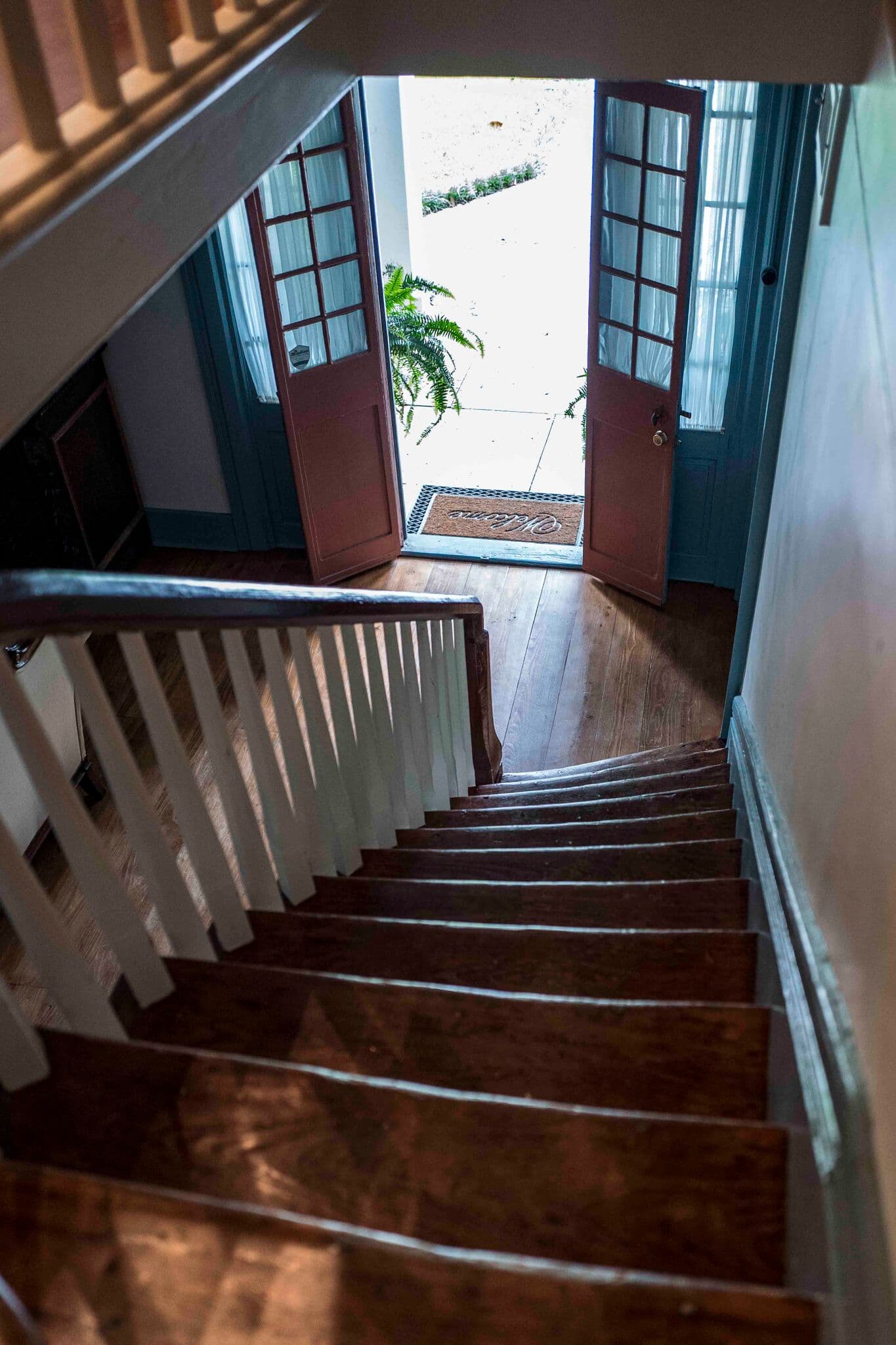 A staircase leads down to open double doors, revealing a light-filled entrance with a doormat and potted plants outside.