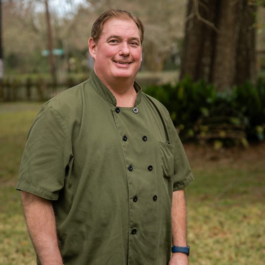 A smiling man wearing a green chef's coat stands outdoors in a grassy area.