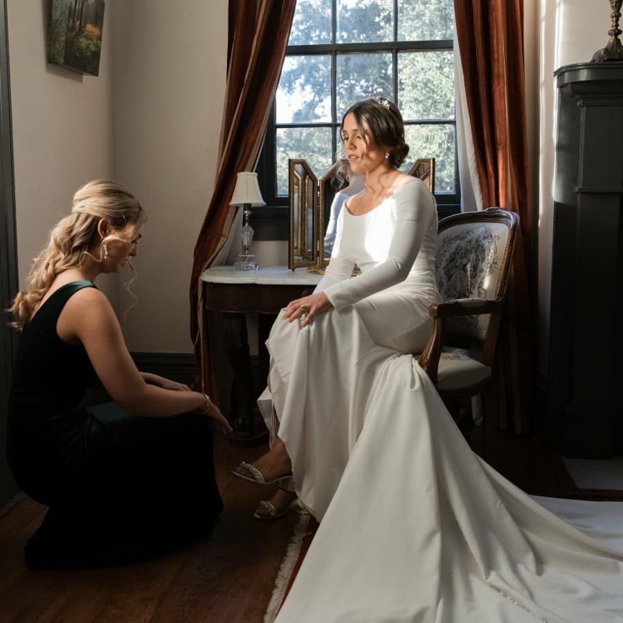 A bride in a white dress sits in a chair while a friend kneels beside her in a dimly lit room.