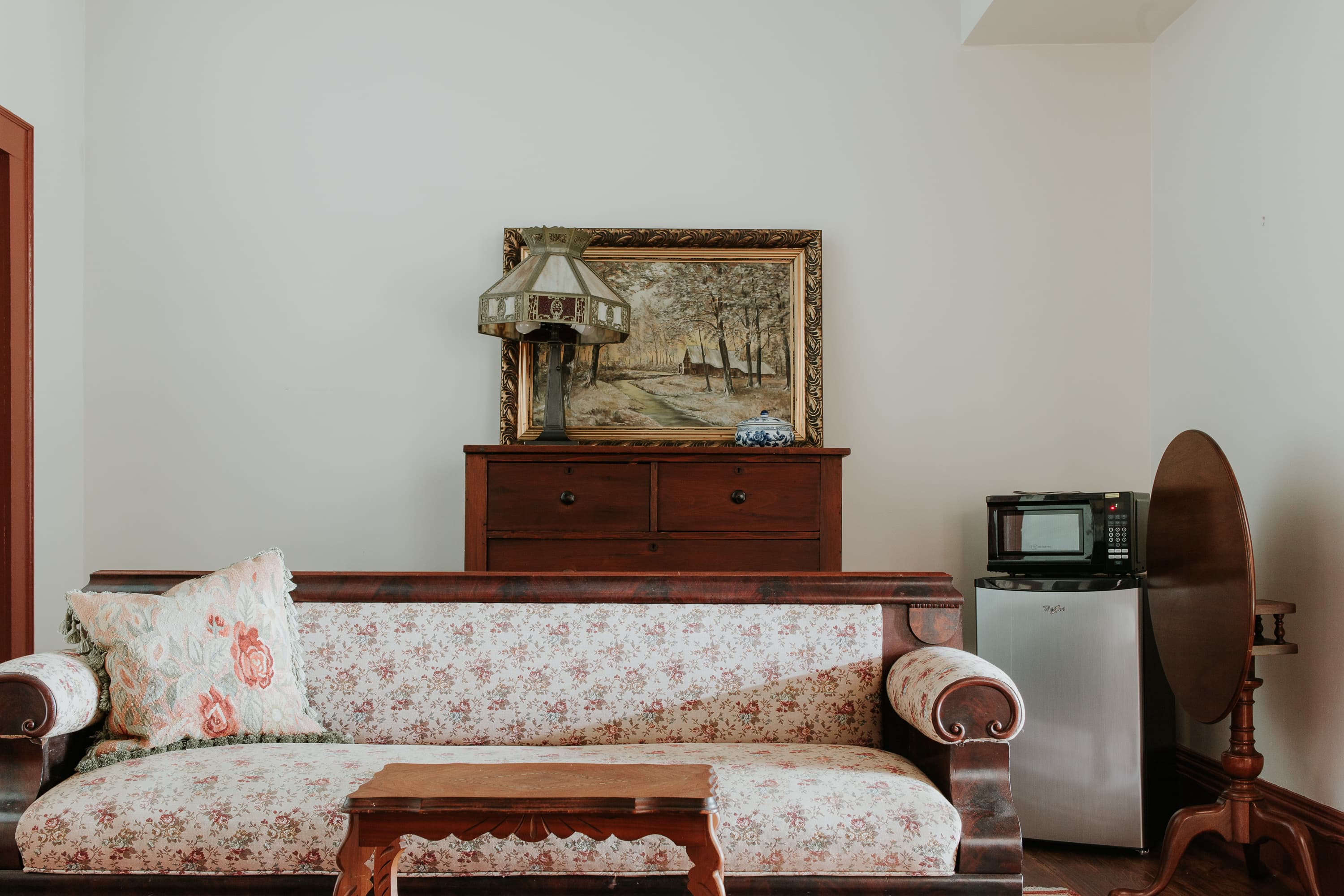 A vintage living room with a floral sofa, a small wooden table, a framed landscape painting, and a microwave next to a mini refrigerator.