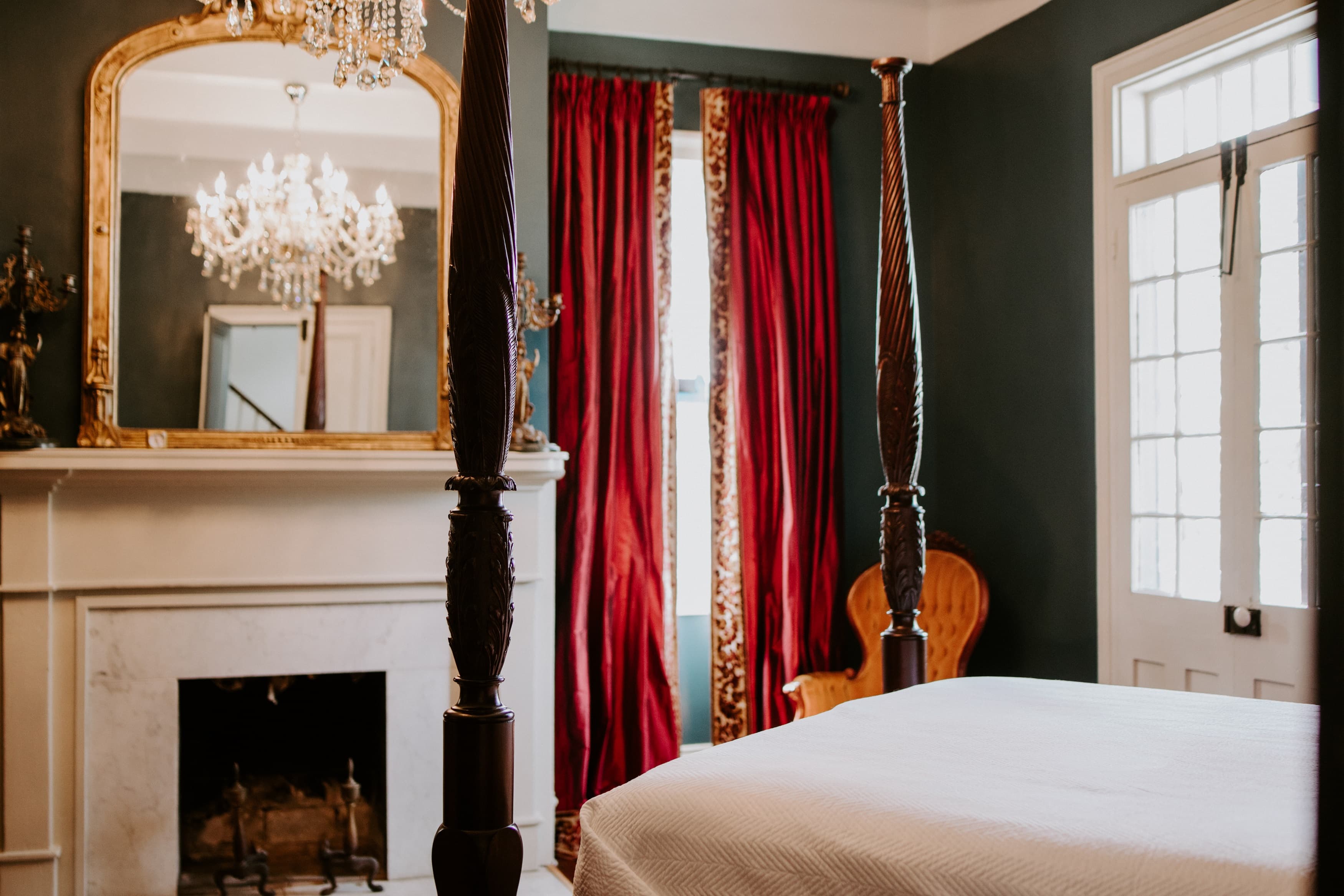 A cozy bedroom featuring a four-poster bed, ornate mirror, red curtains, and a vintage armchair.