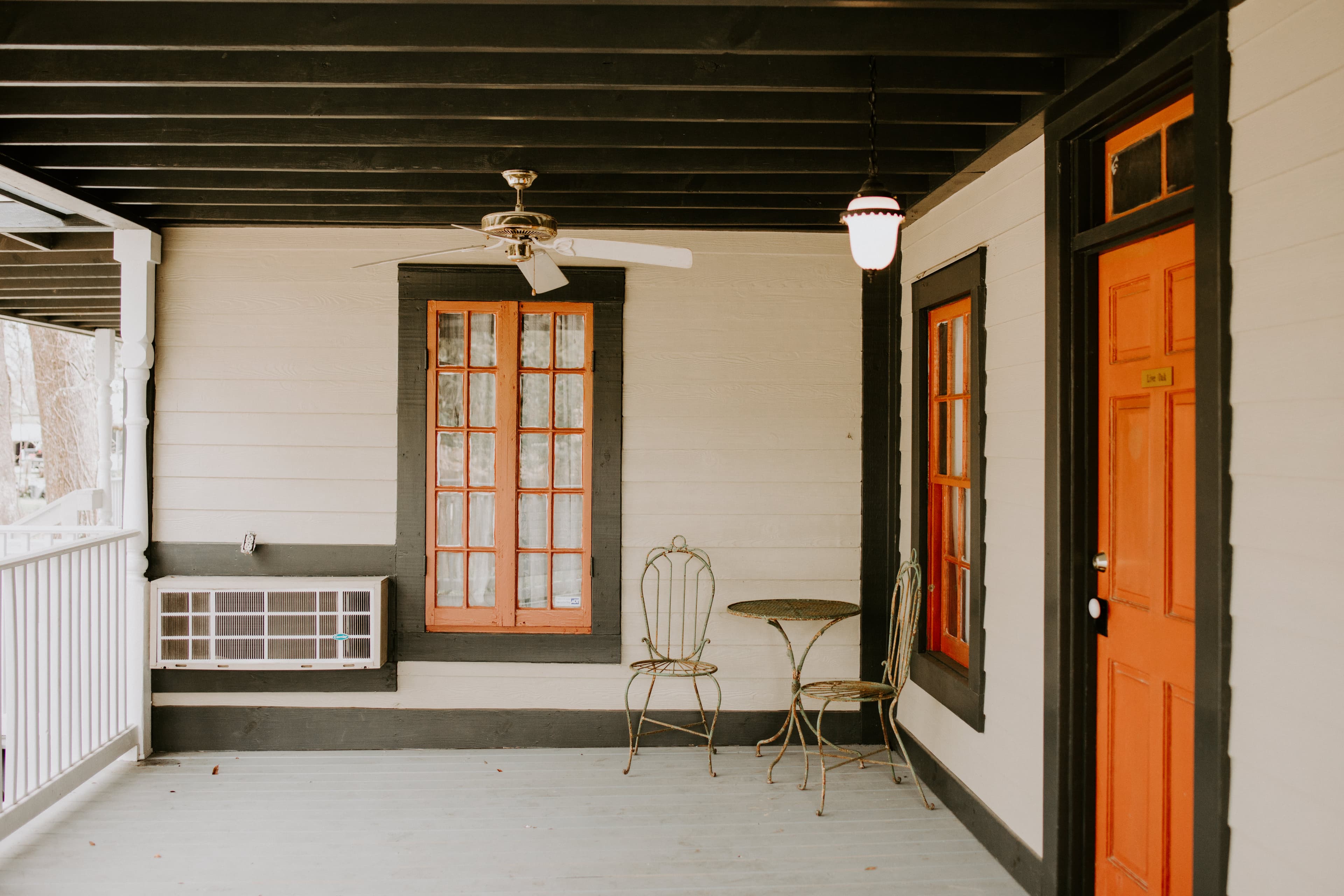 A cozy porch featuring orange-trimmed windows and door, a small table with chairs, and an air conditioning unit.