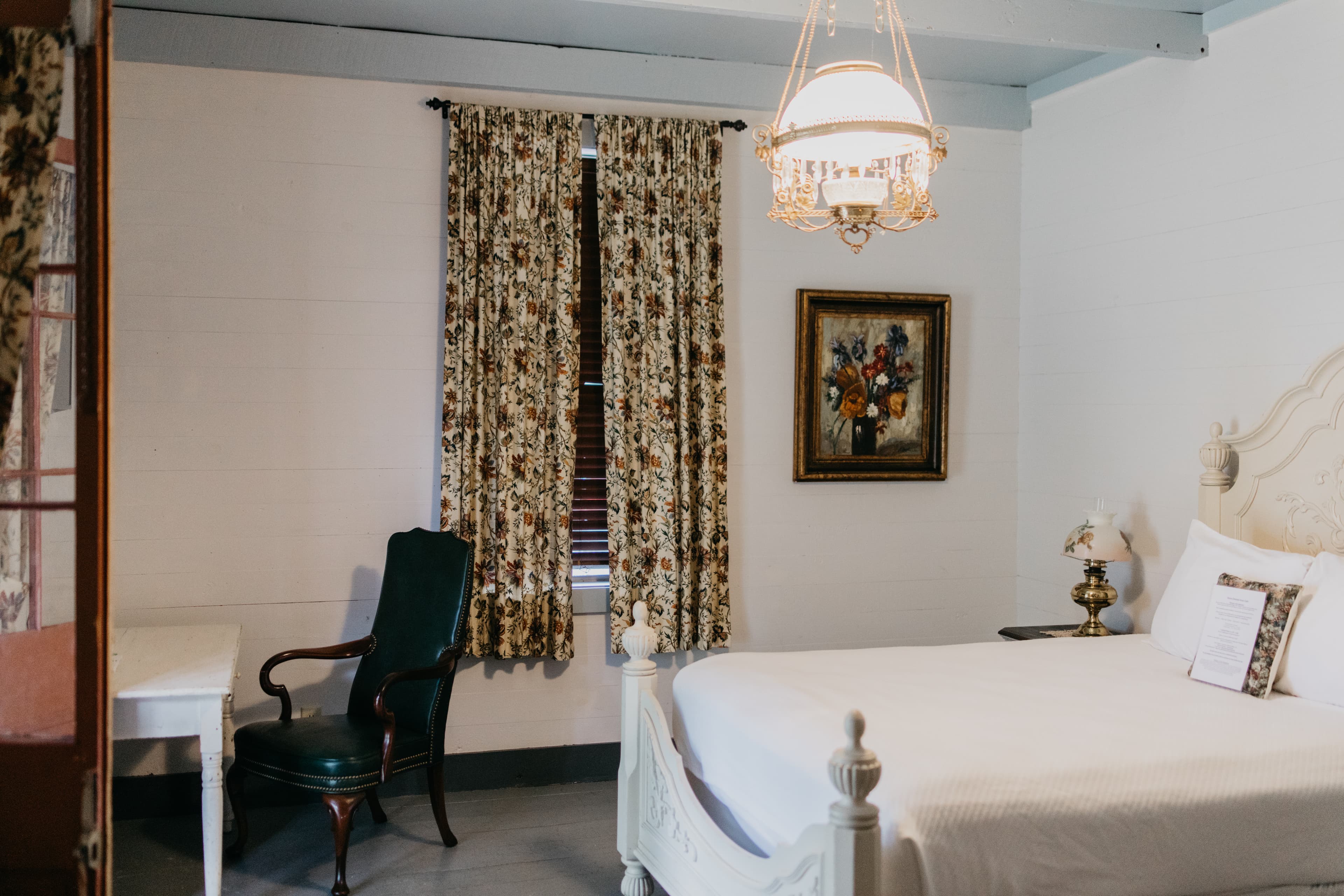 A cozy bedroom featuring a white bed, floral curtains, a green chair, and a vintage lamp.
