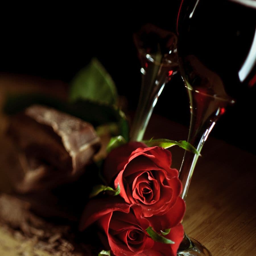 Two glasses of red wine beside red roses and a piece of chocolate on a wooden surface.