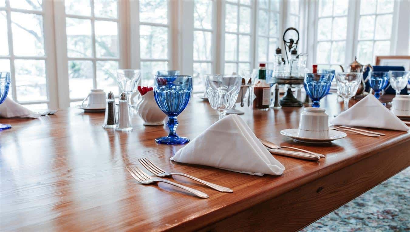 A dining table elegantly set with blue glassware, folded napkins, and fine cutlery beside large windows.