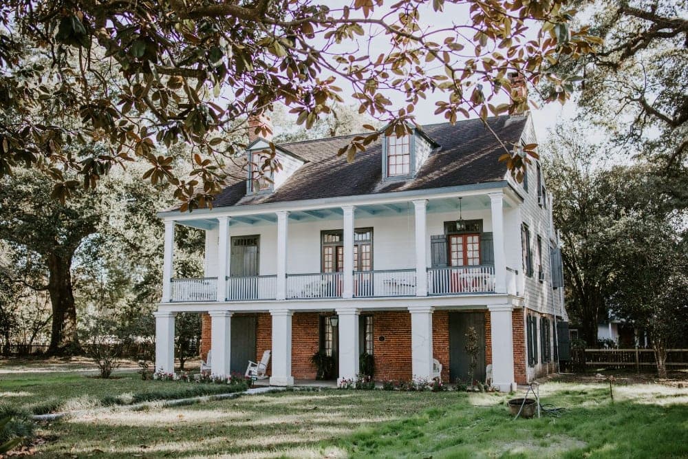 A historic two-story house with a wraparound porch, surrounded by greenery.
