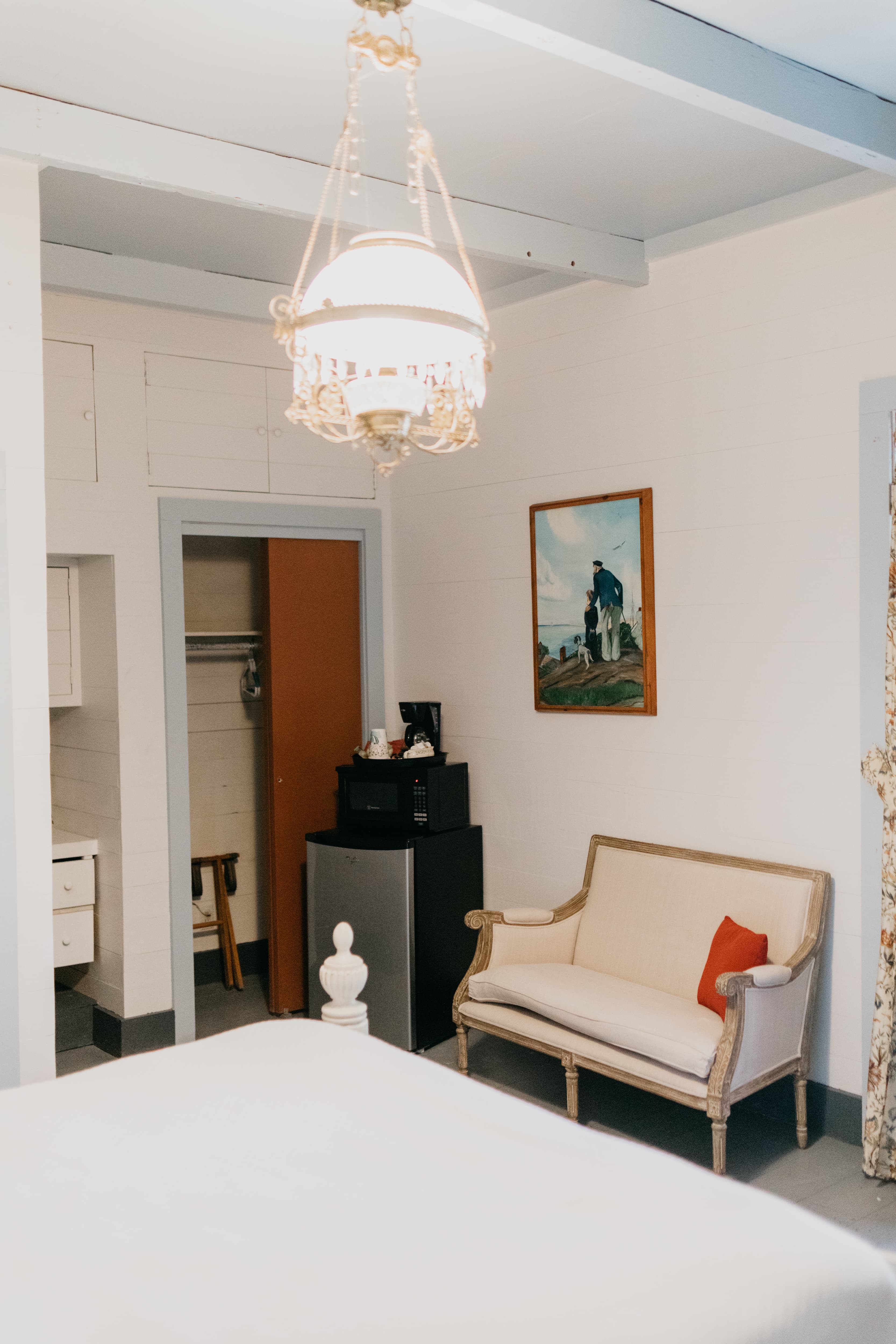 A cozy room featuring a vintage chandelier, a small couch, a microwave, and a framed painting on the wall.