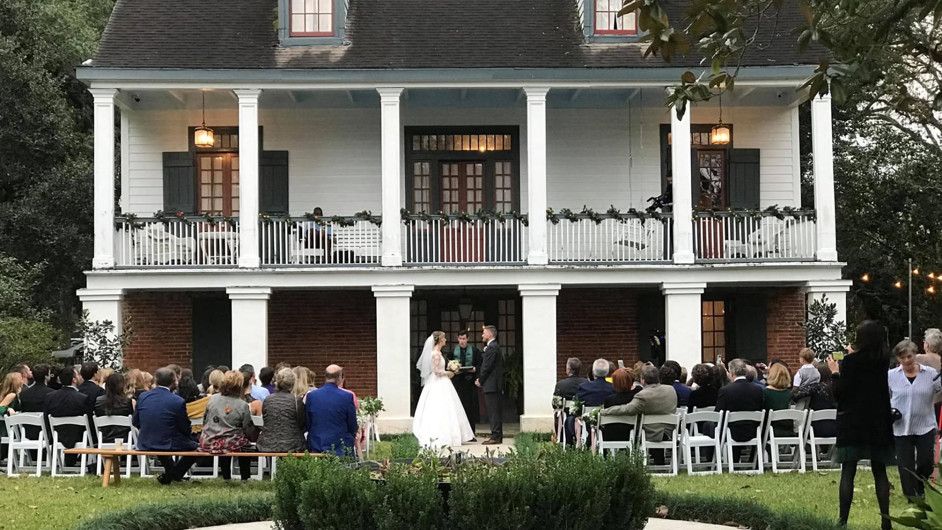 A couple exchanges vows during an outdoor wedding ceremony in front of a historic house, surrounded by guests.