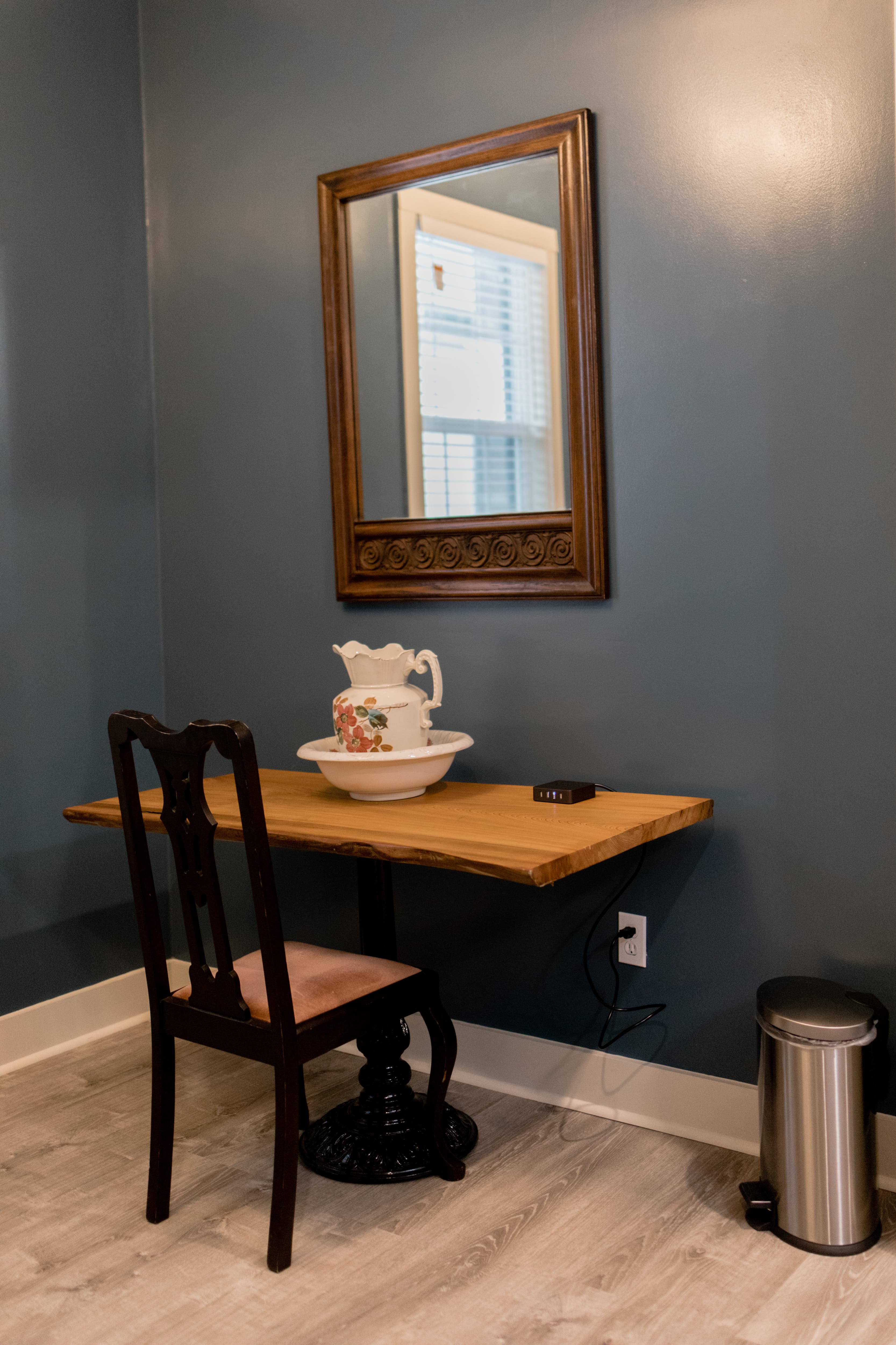 A small table with a pitcher and bowl, a vintage chair, and a mirror against a blue wall.