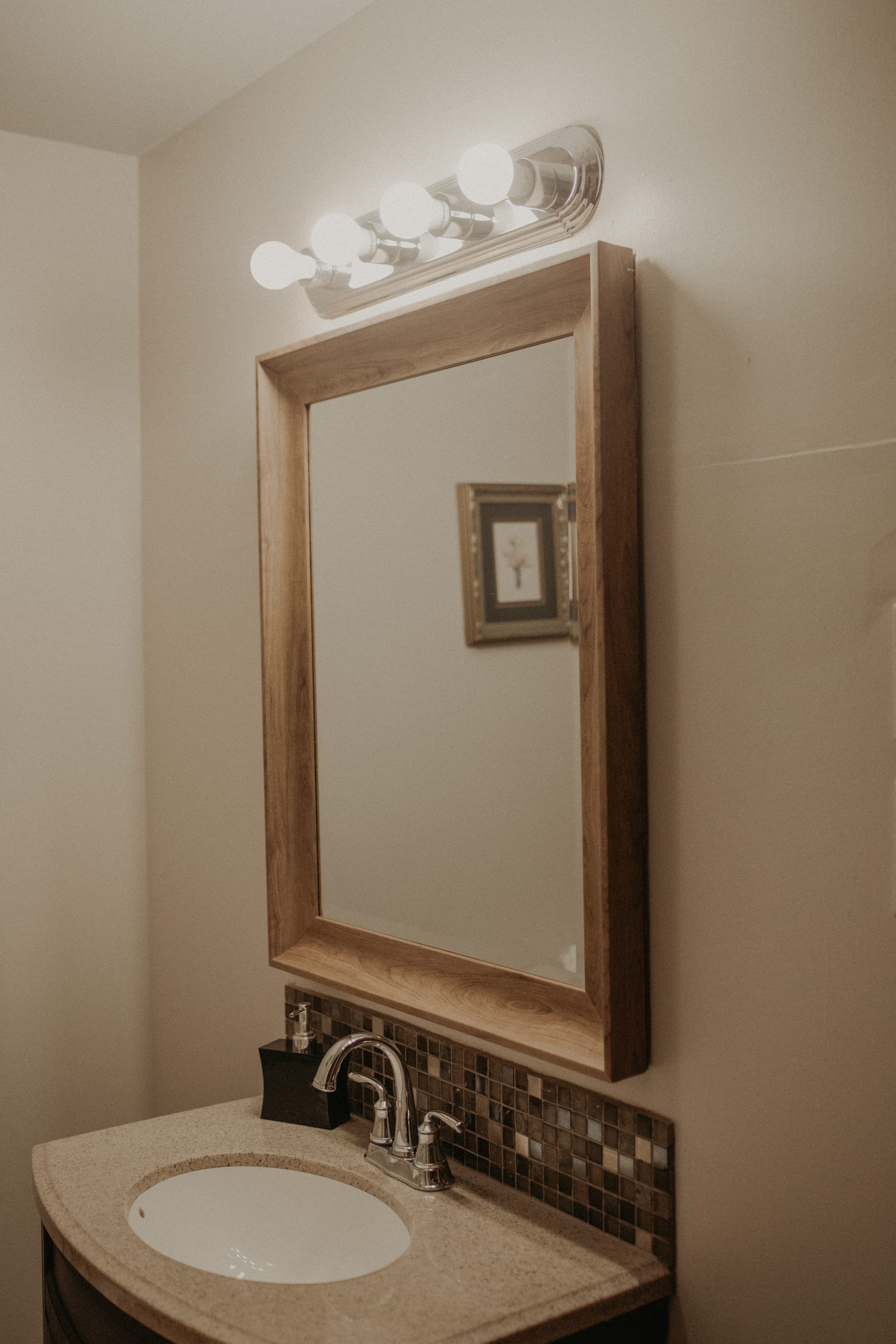 A well-lit bathroom vanity with a wooden-framed mirror, sink, and decorative tile.