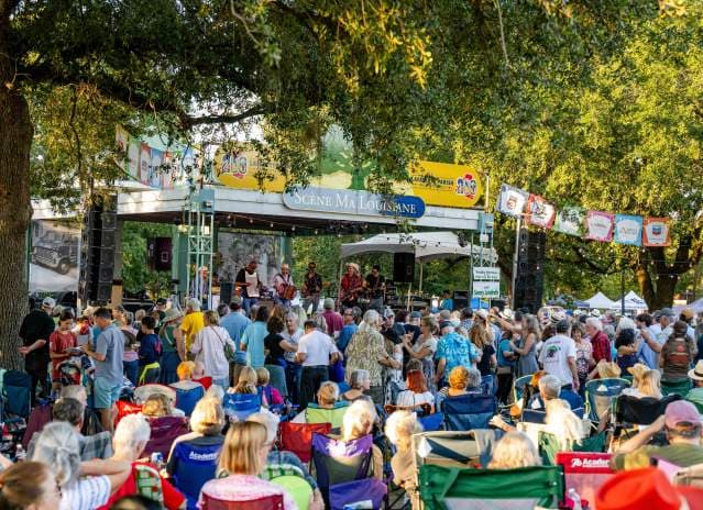 A large crowd enjoys a live music performance at an outdoor festival under shady trees.