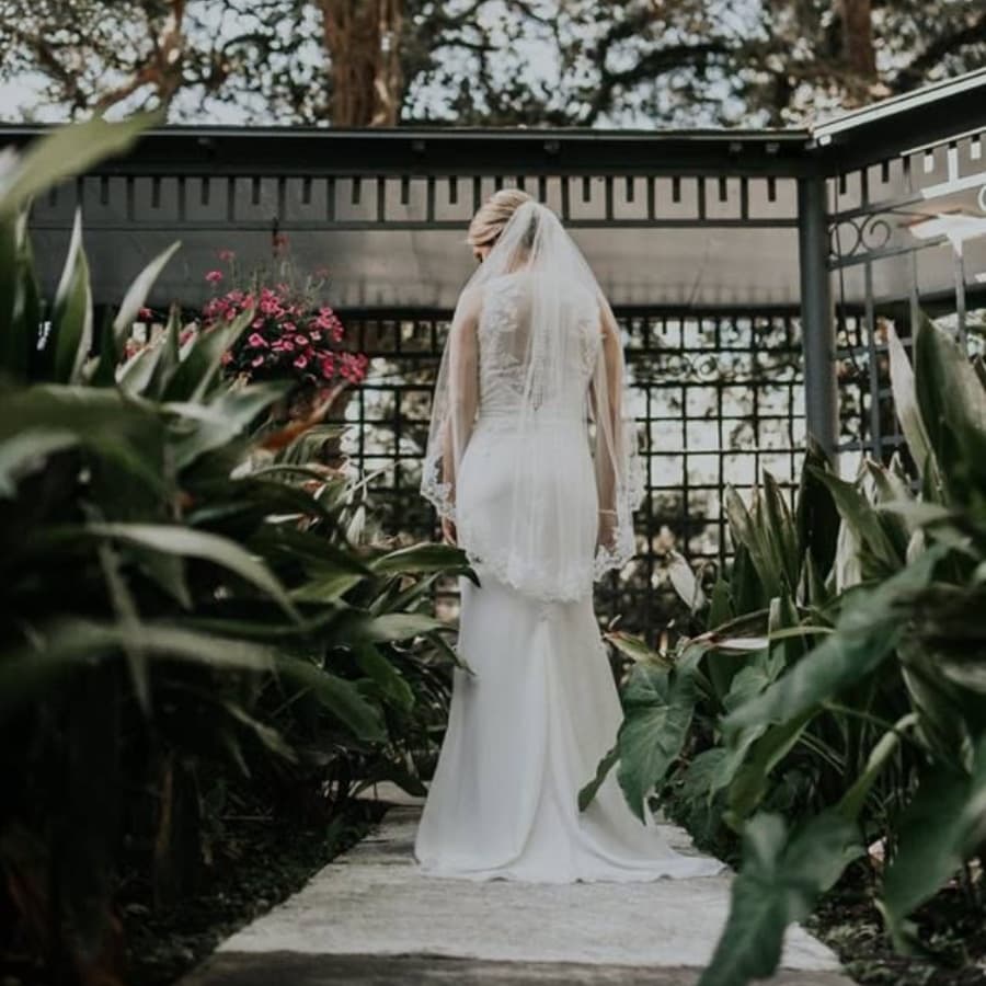 A bride in a white wedding gown walks along a stone path surrounded by lush greenery and flowers.