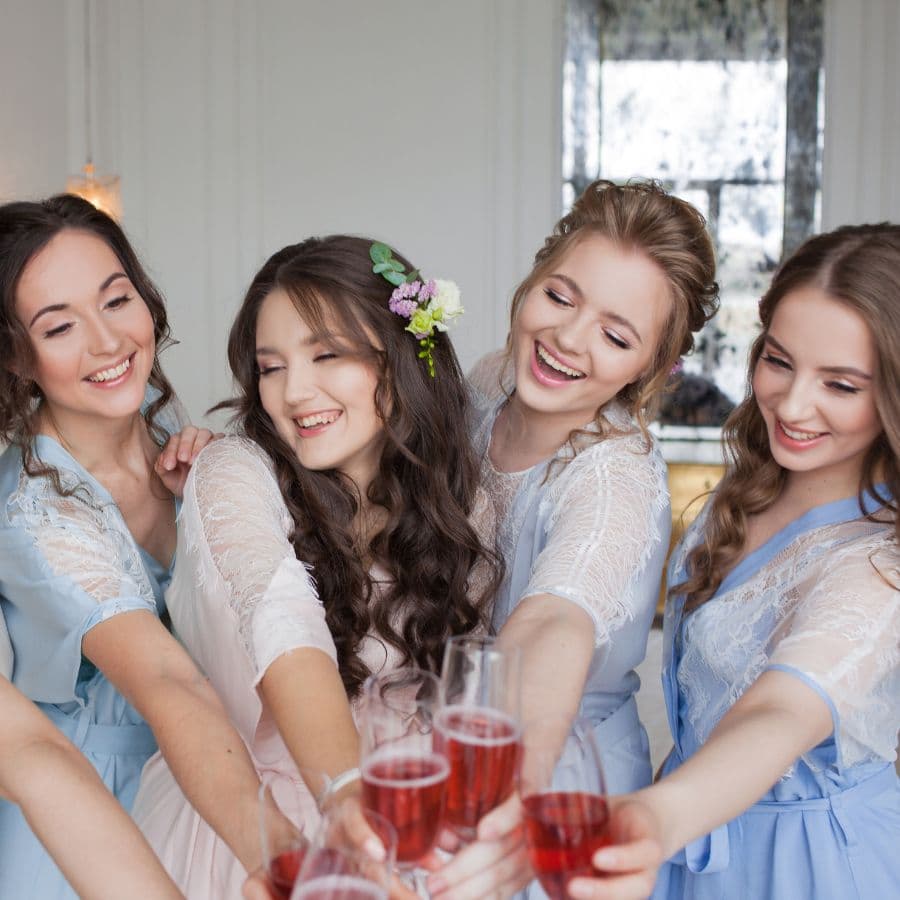 A group of four joyful women in pastel dresses cheers with glasses of pink drinks.