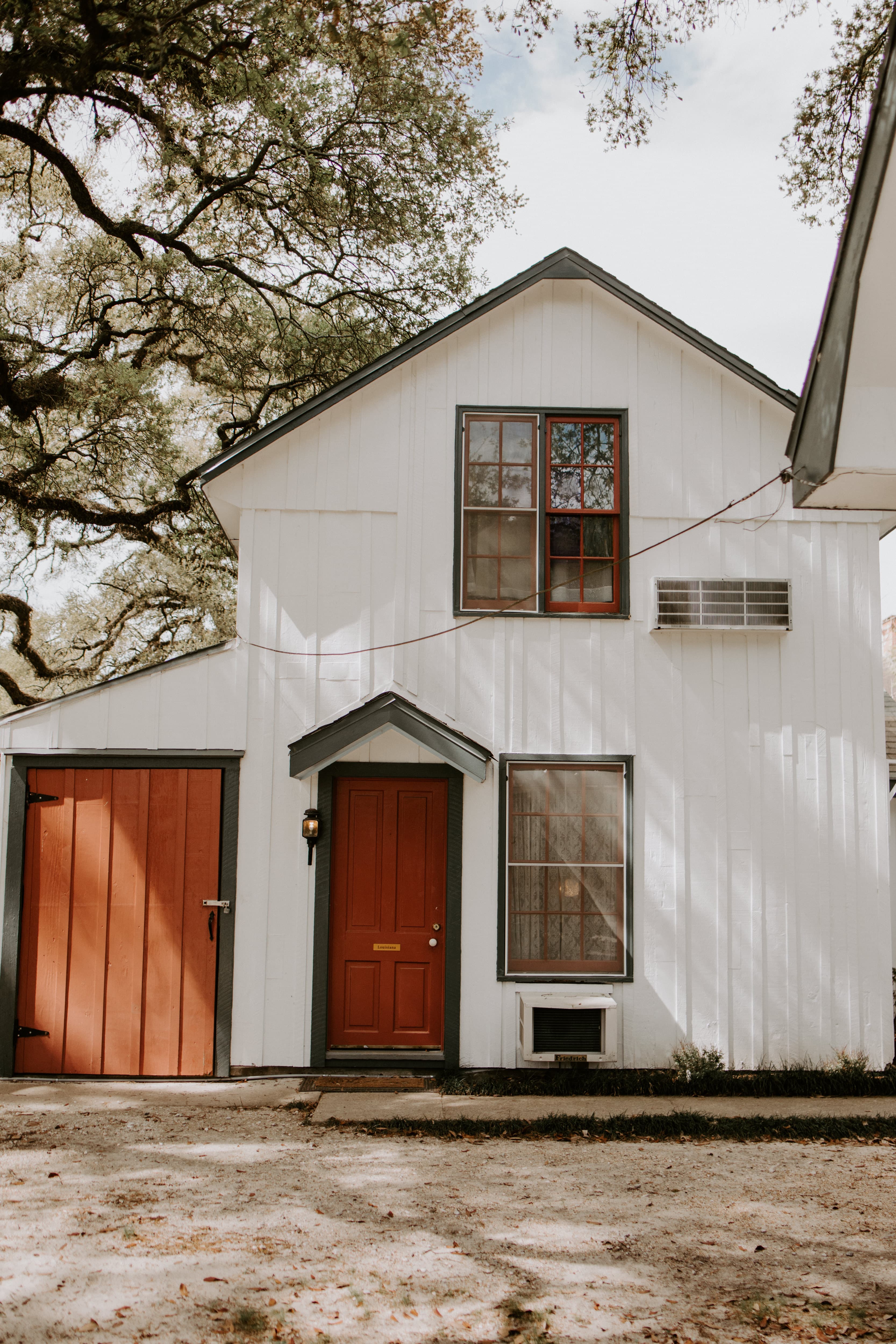 A white, two-story house with a red door and matching garage door, surrounded by trees.