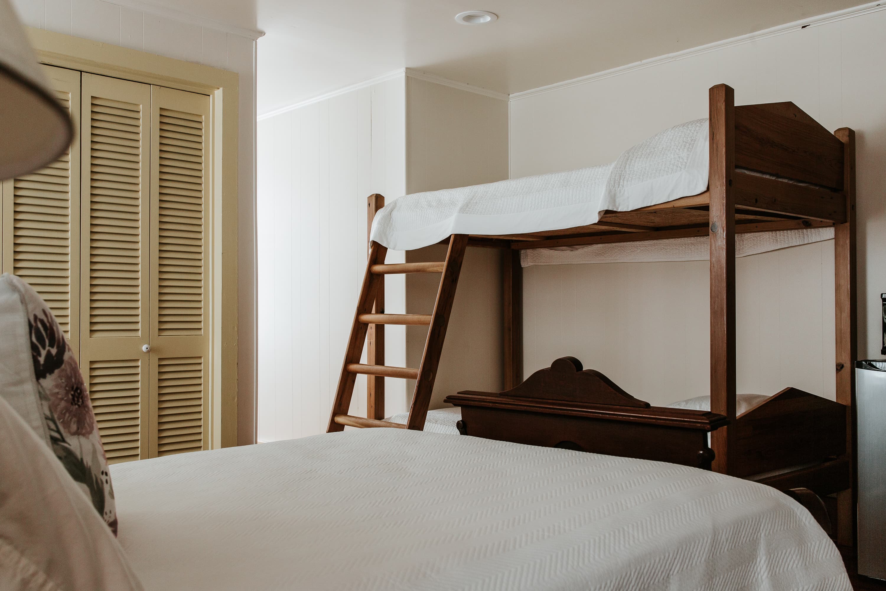 A wooden bunk bed with a ladder next to a double bed in a simple, light-colored room.