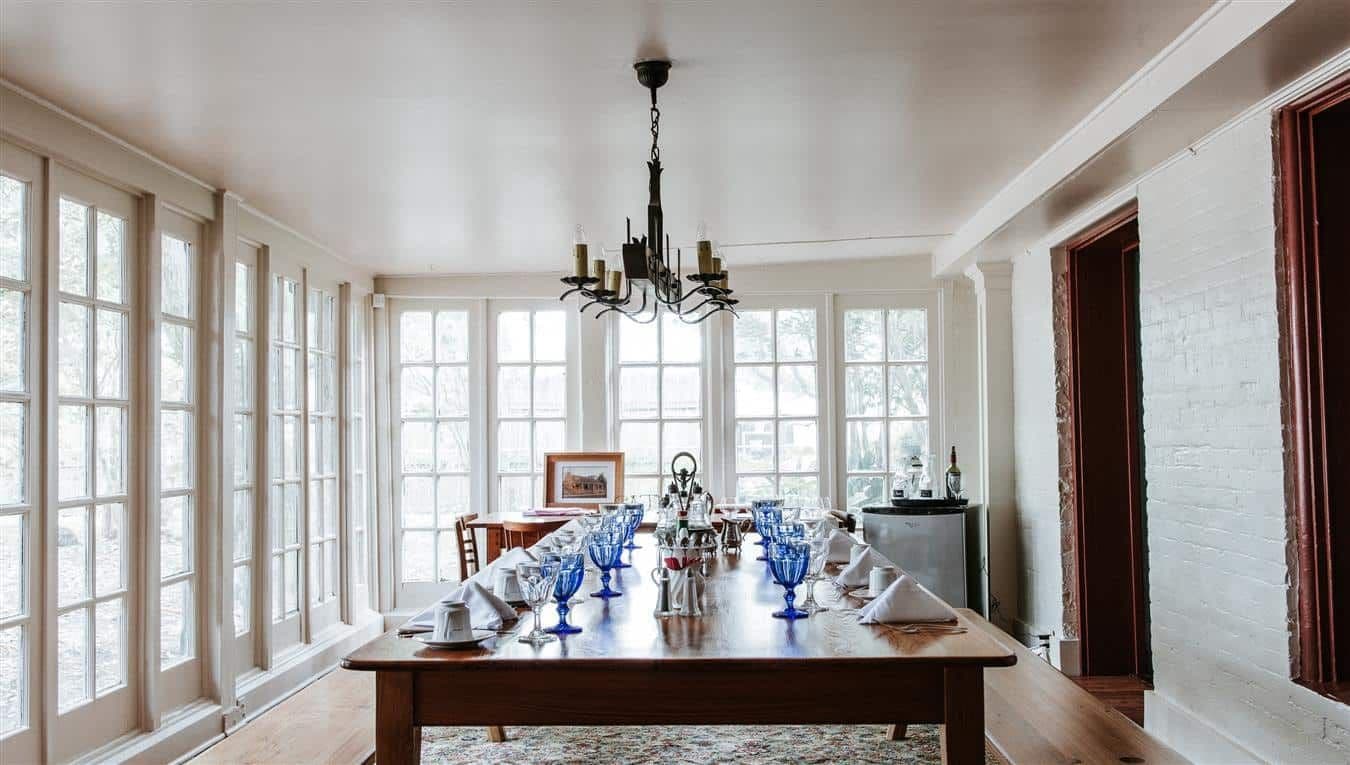 A brightly lit dining room featuring a large wooden table set with blue glasses and white napkins, surrounded by large windows.