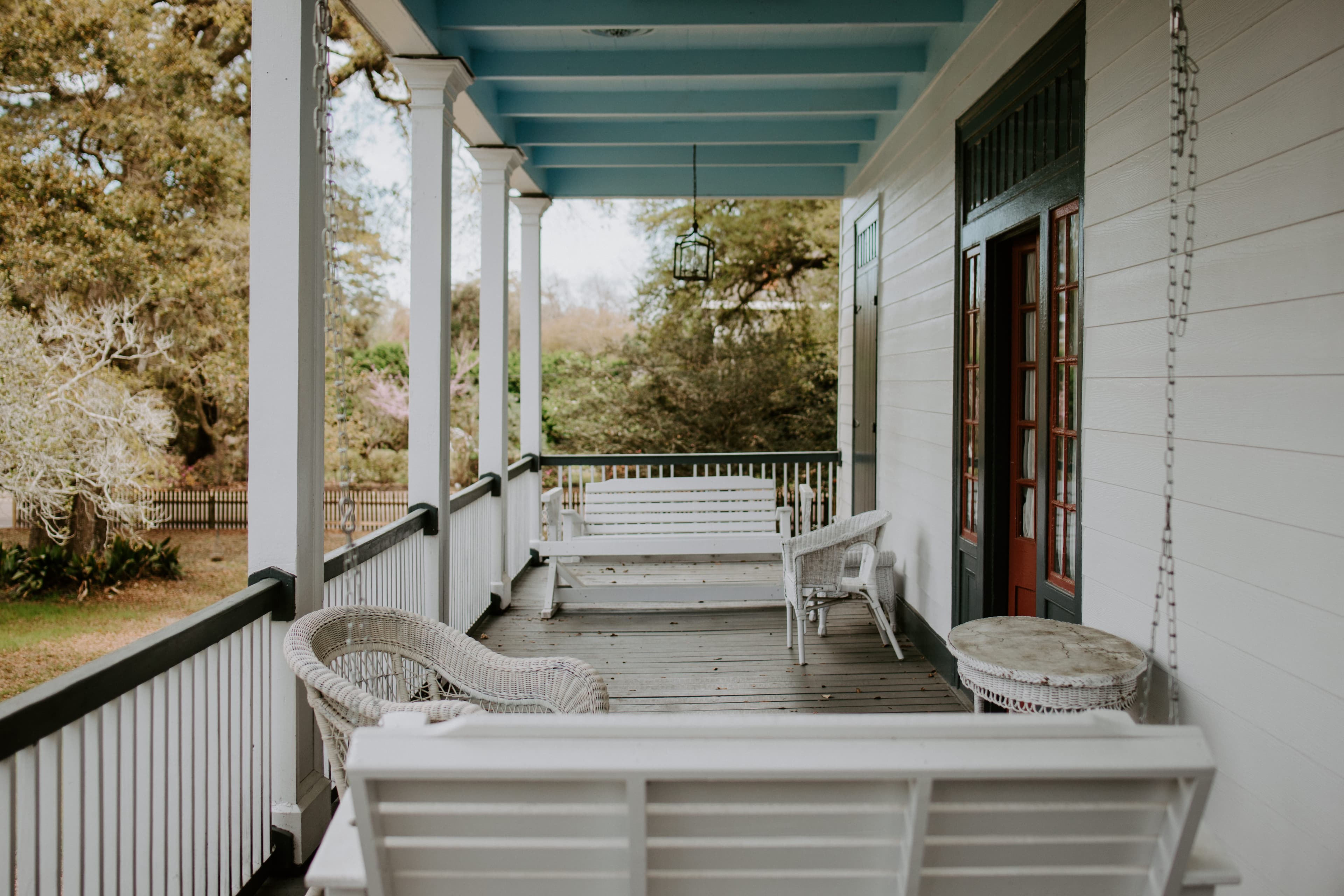 A charming porch featuring white wicker furniture and a hanging lantern, surrounded by greenery.