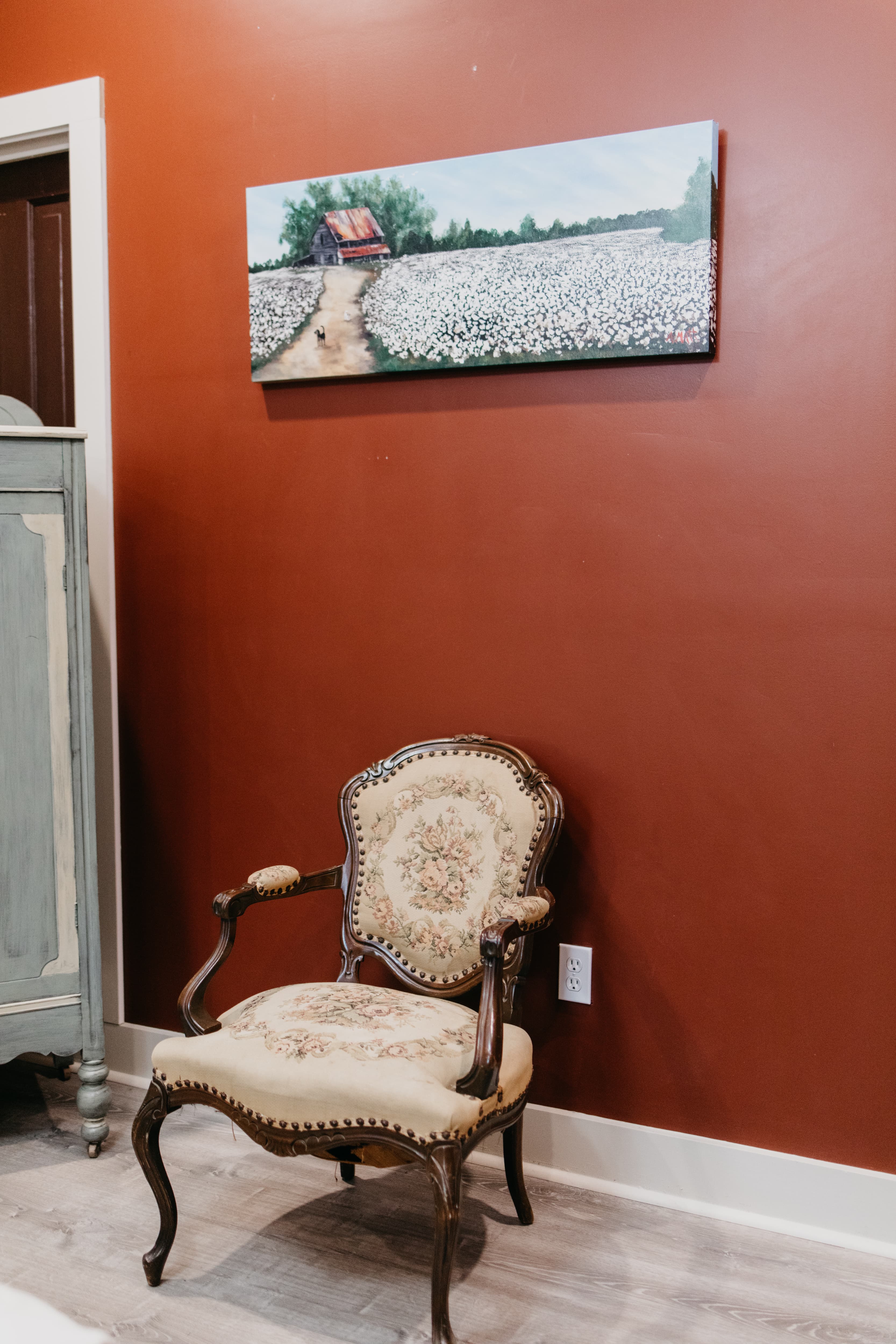 A vintage upholstered chair sits against a rust-colored wall, beneath a landscape painting of a cotton field.