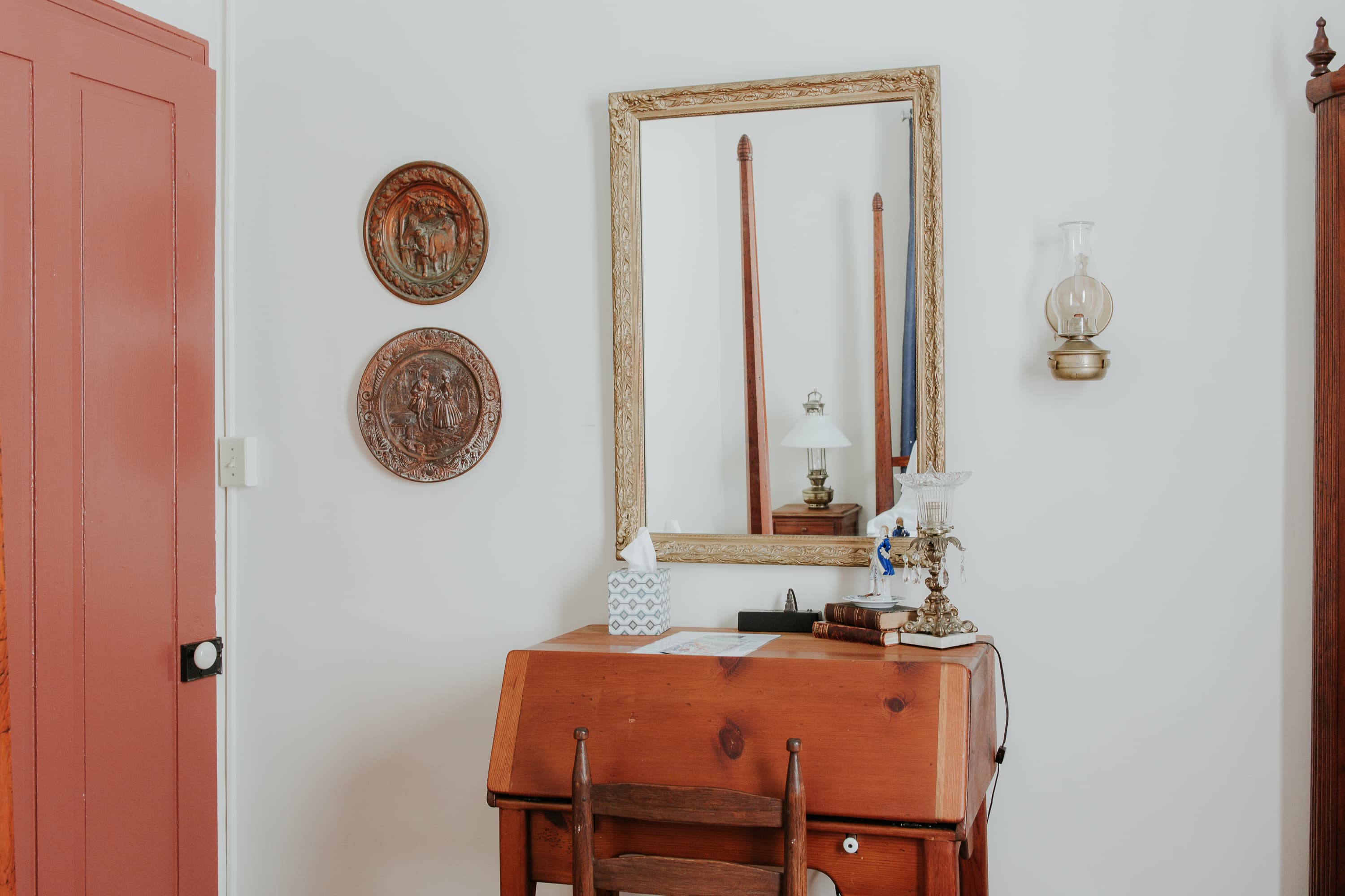 A wooden writing desk with a mirror and decorative wall plates in a softly lit room.