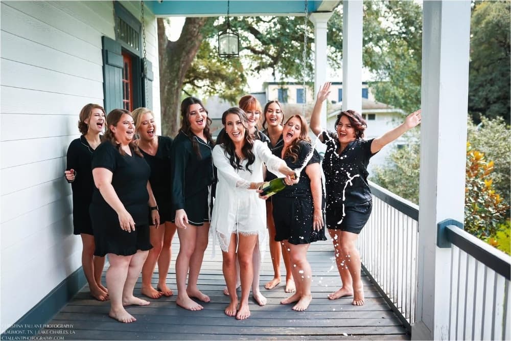 A group of joyful women in black outfits celebrate together, with one woman popping a champagne bottle on a porch.