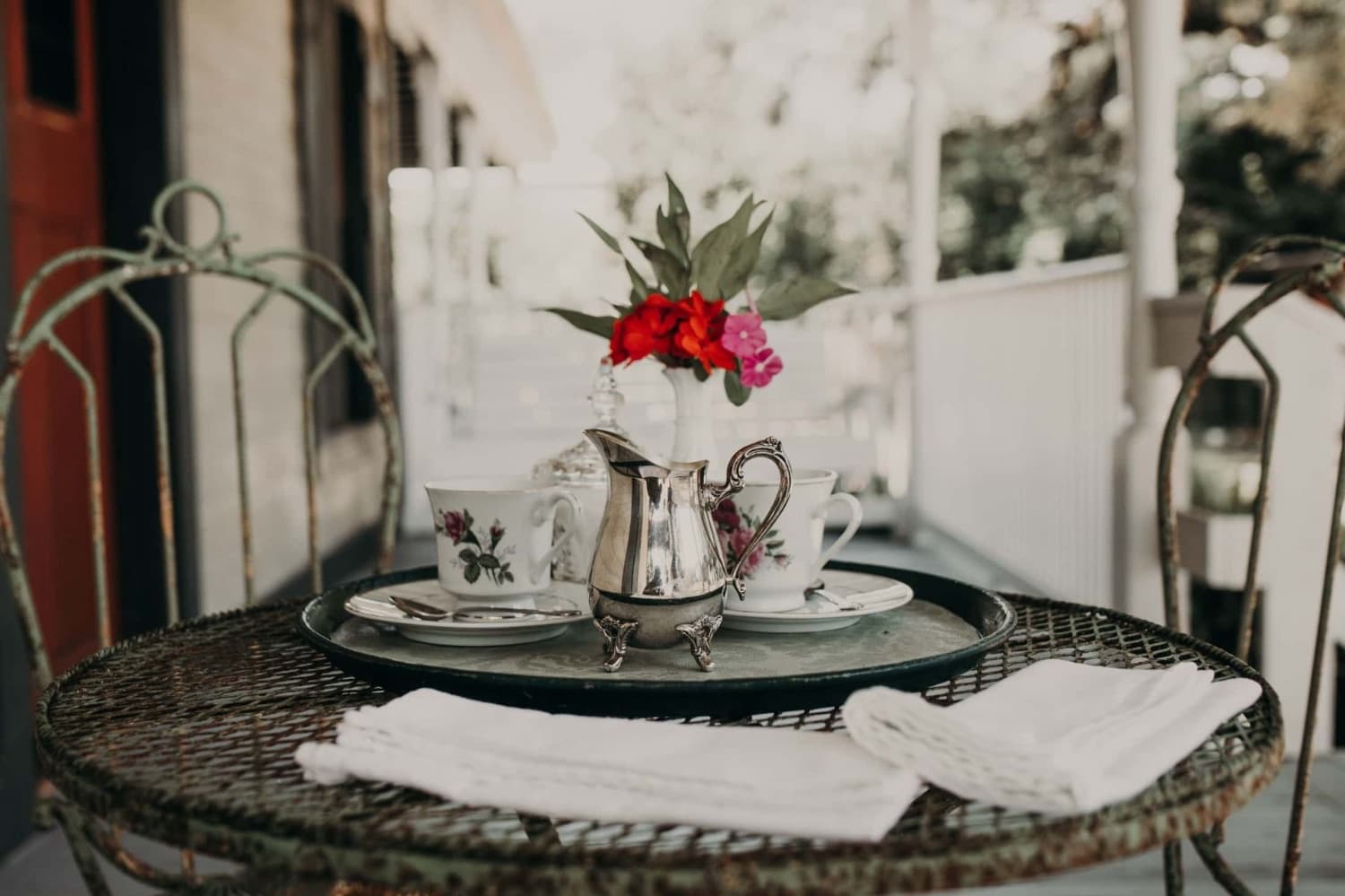 A vintage tea set with a silver pitcher and floral cups sits on a rustic table adorned with a bouquet of flowers.