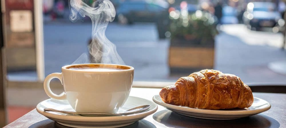 A steaming cup of coffee alongside a buttery croissant on a table with a bright window view.
