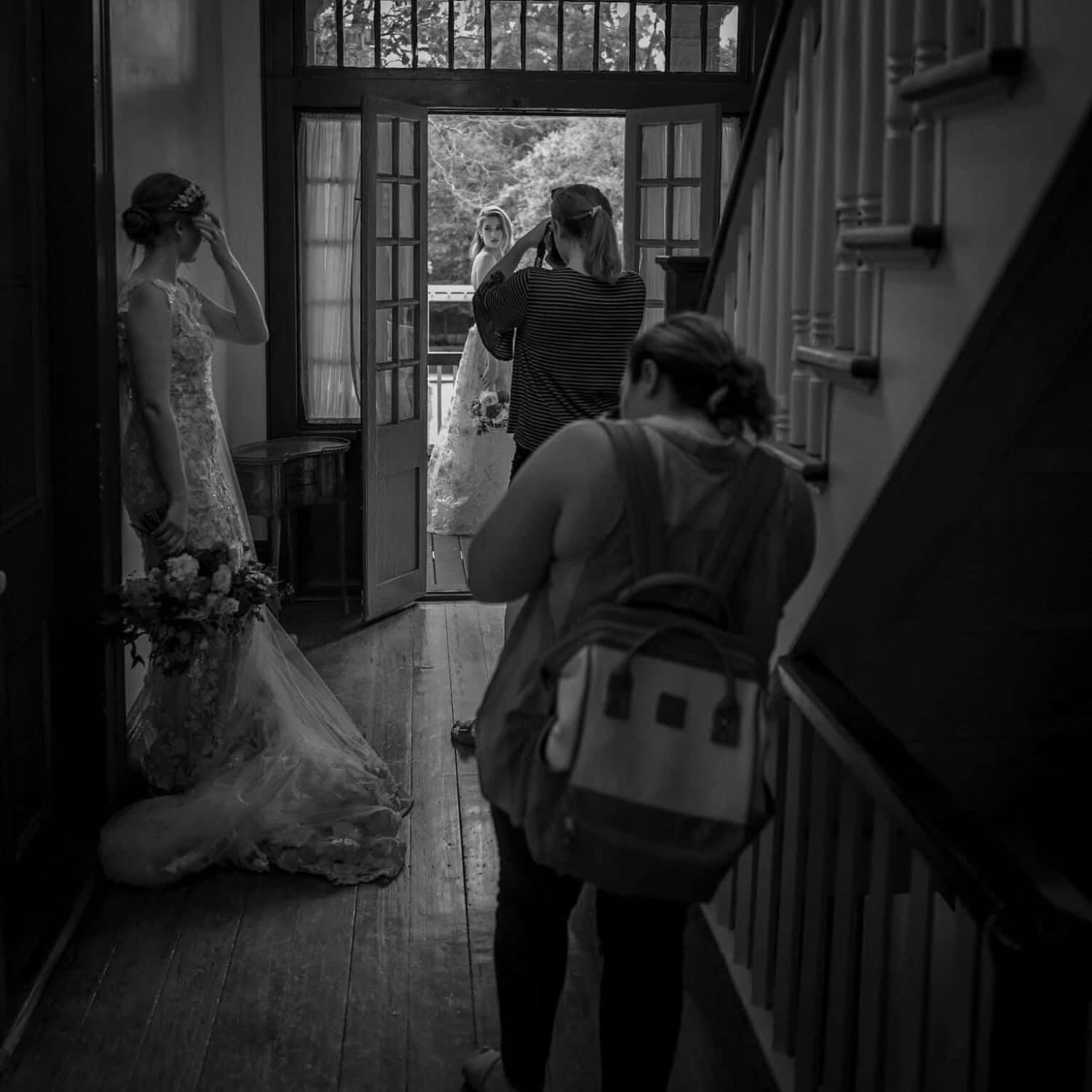 A bride in a wedding dress stands at the doorway while a photographer captures the moment with two other women present in the hallway.