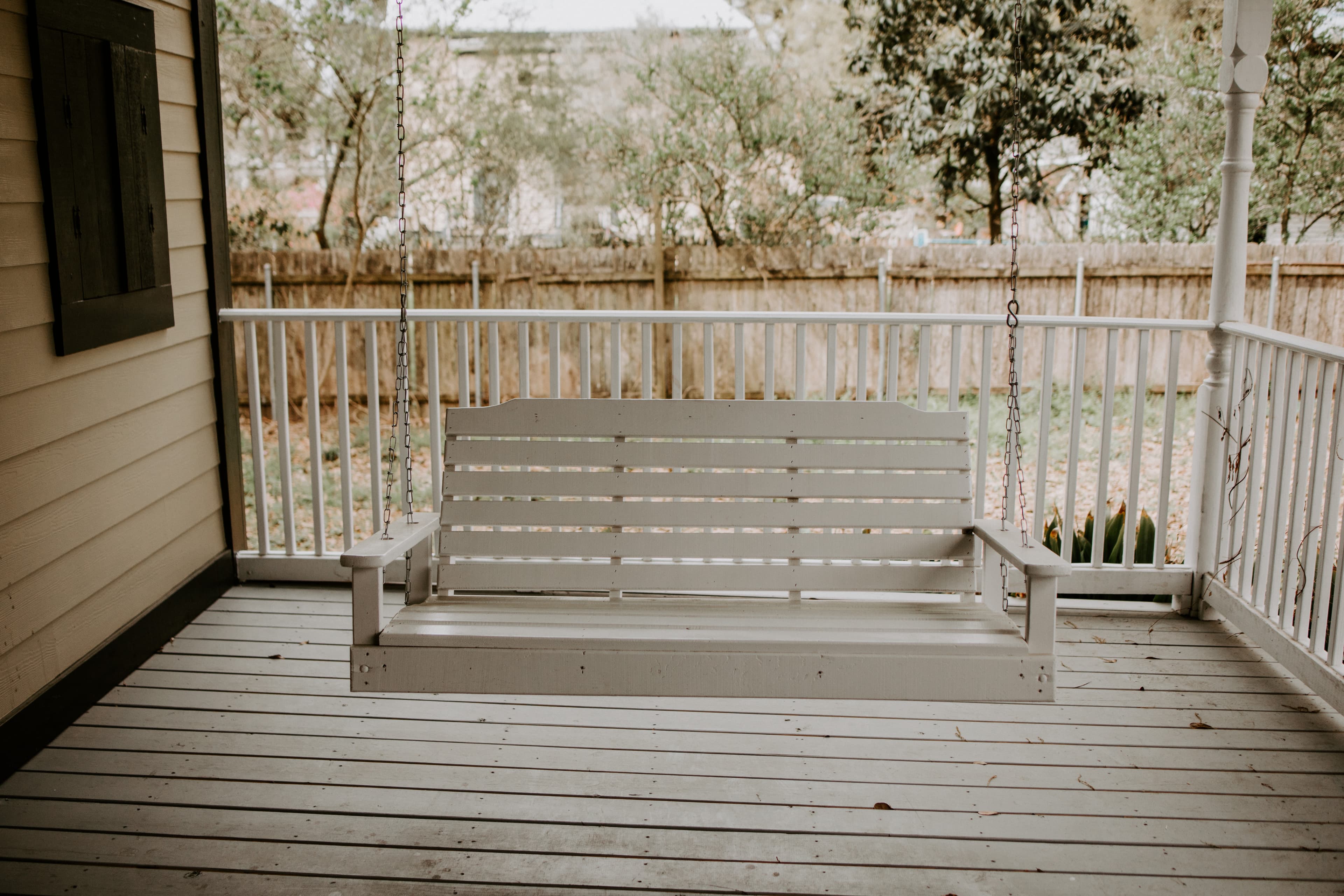 A white wooden porch swing is suspended on a covered porch with a fenced yard in the background.