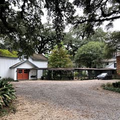 A gravel driveway leads to a rustic house with orange doors and a covered parking area, surrounded by lush greenery.