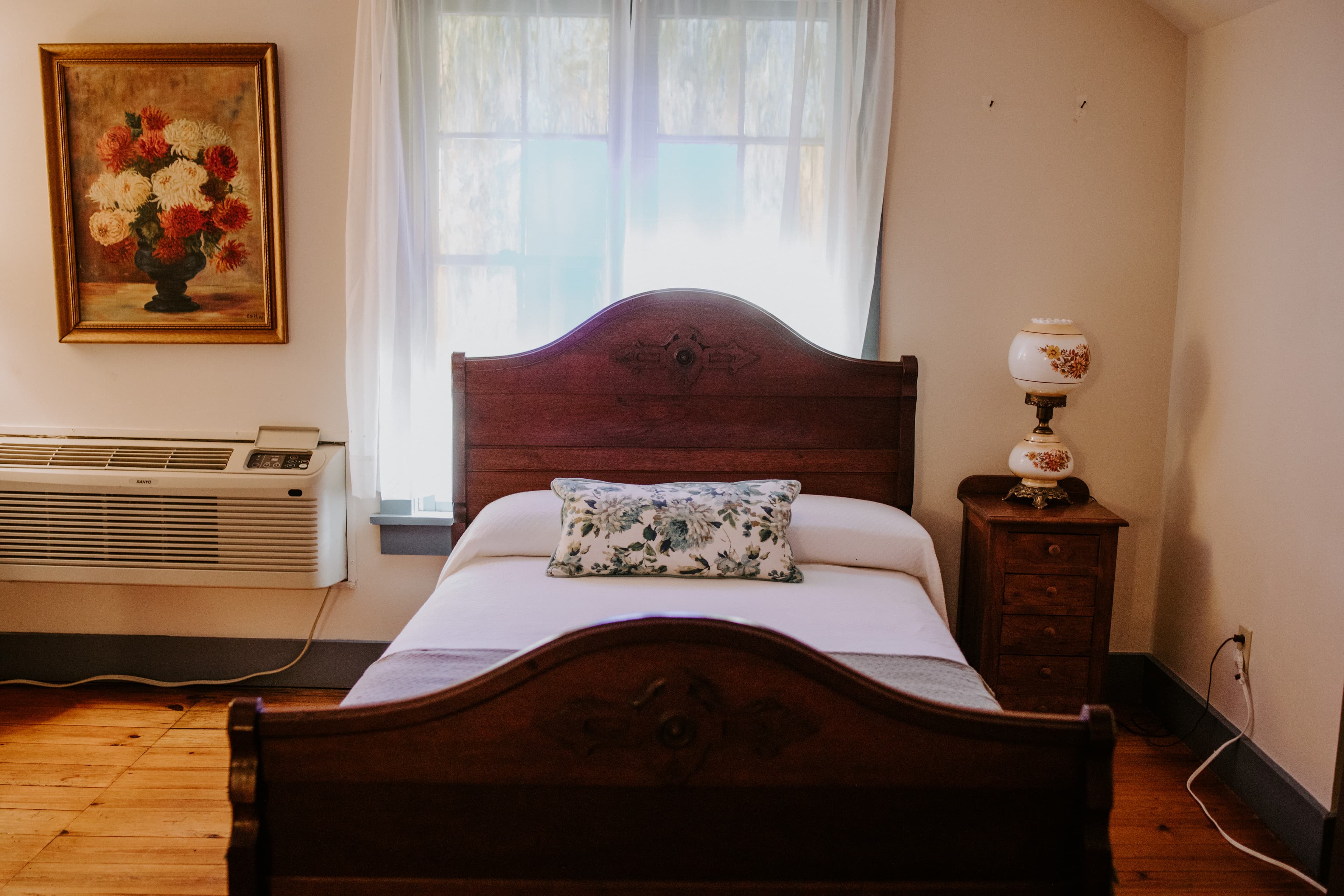 Cozy bedroom featuring a wooden bed, decorative pillow, vintage lamp, air conditioner, and a floral painting on the wall.