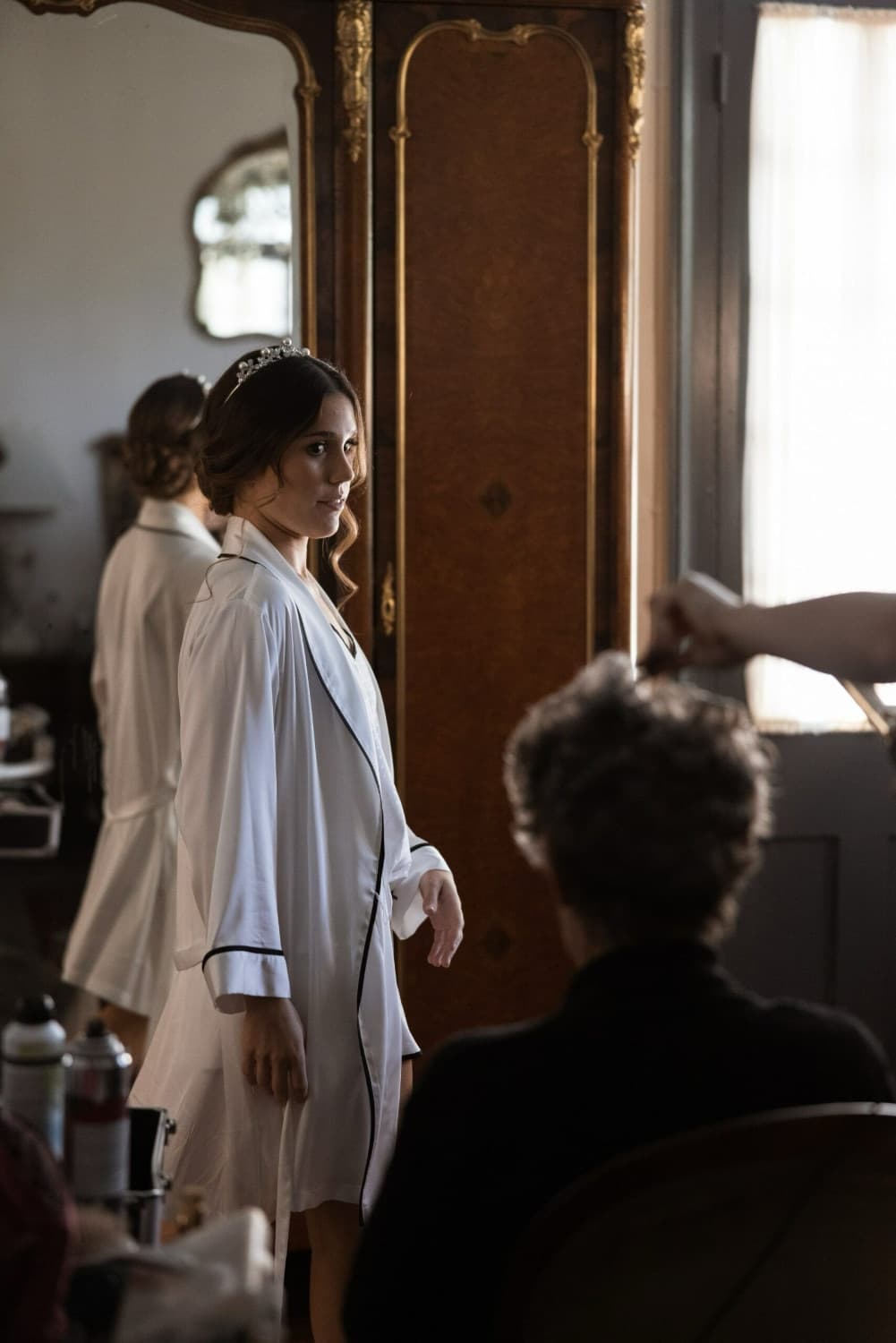 A woman in a white robe and tiara poses in front of a mirror while getting ready, with another person seated in the foreground.