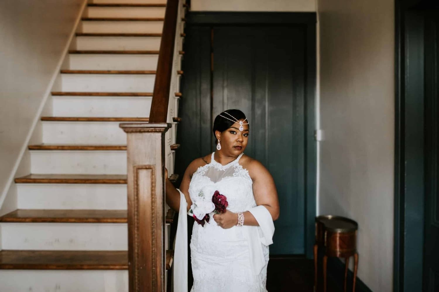 A bride in a white wedding dress stands by a staircase, holding a bouquet and looking confidently at the camera.