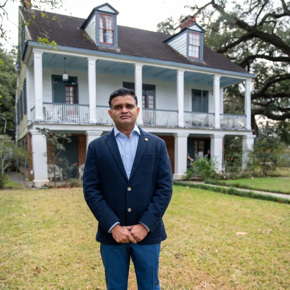 A man in a dark blazer stands in front of a historic white house with blue accents.