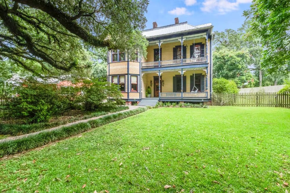 A large, colorful two-story house with a porch surrounded by lush greenery and a manicured lawn.