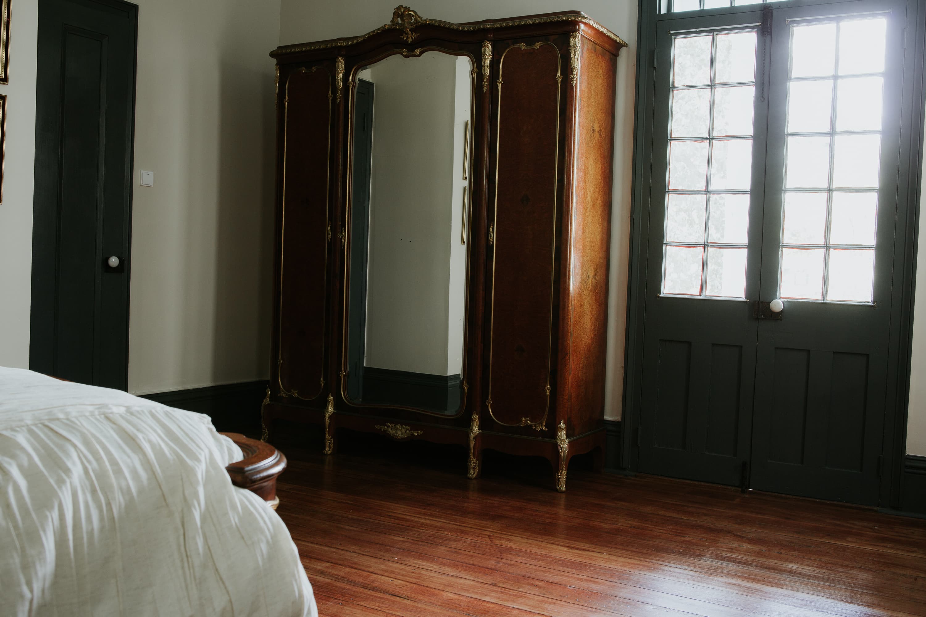 A wooden armoire with a mirror stands in a warmly decorated bedroom beside a large window and a bed.