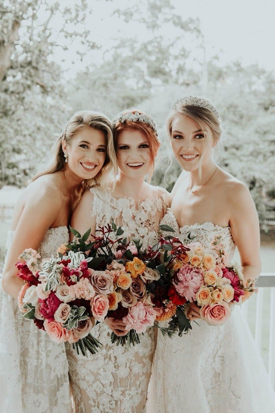 Three smiling bridesmaids in elegant wedding dresses hold colorful bouquets of flowers.