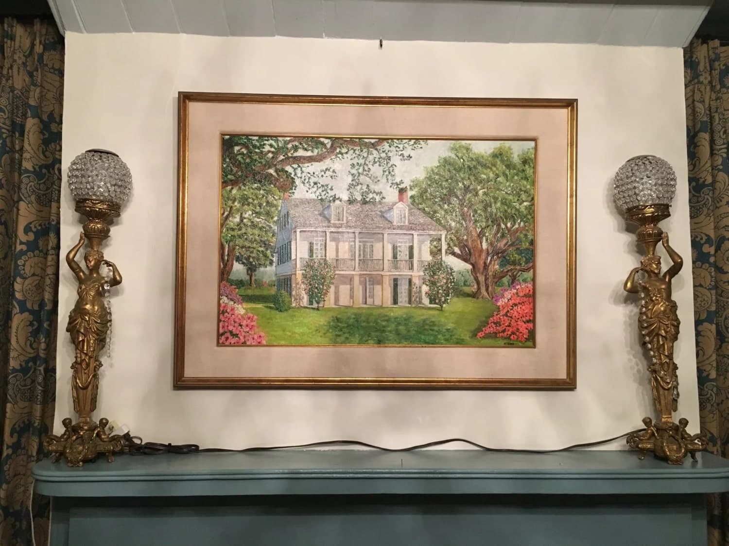 A framed painting of a house surrounded by trees and flowers is displayed above two ornate table lamps.