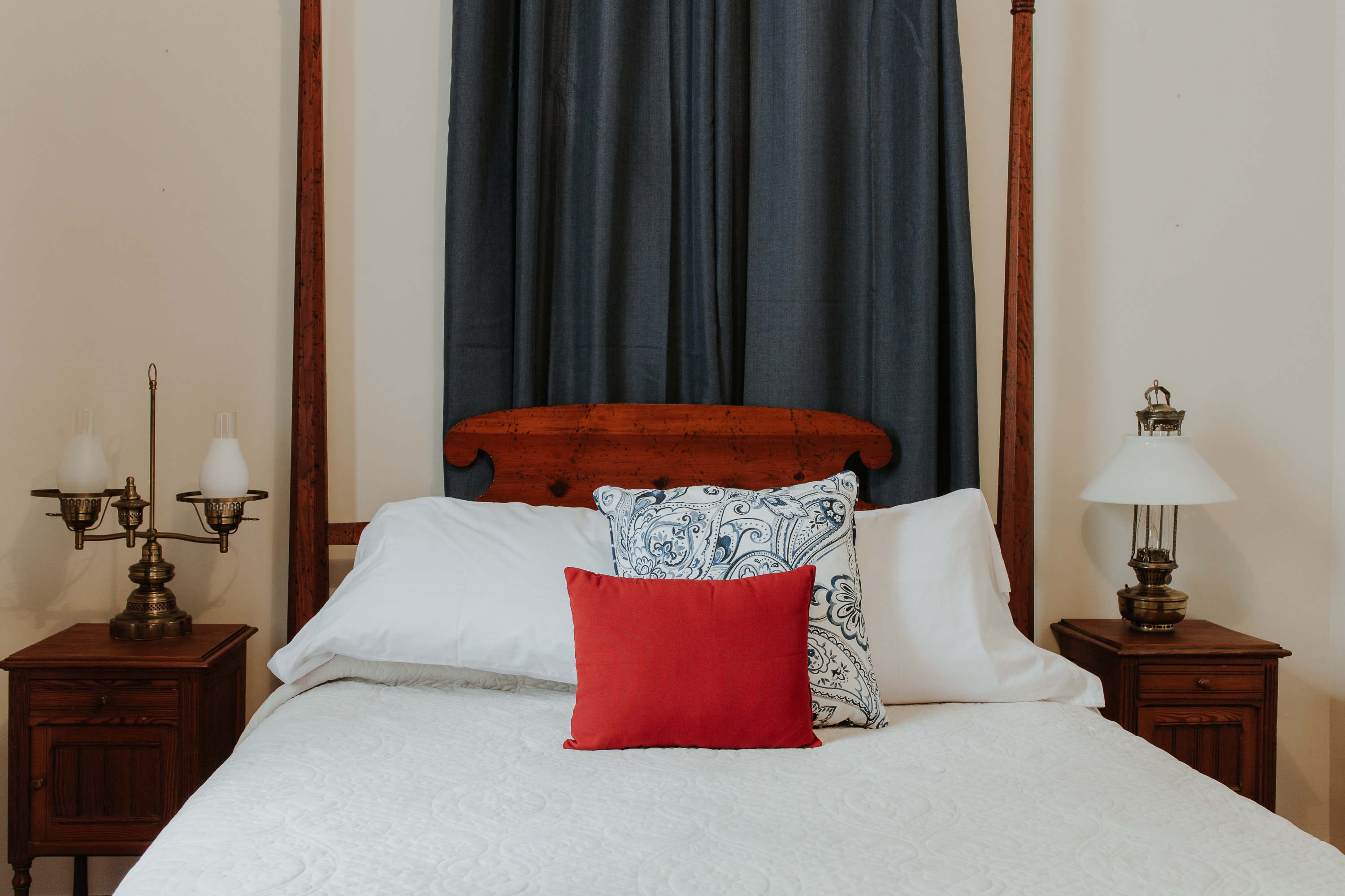 A cozy bed with a white quilt, decorative pillows, and matching bedside lamps against a backdrop of dark curtains.