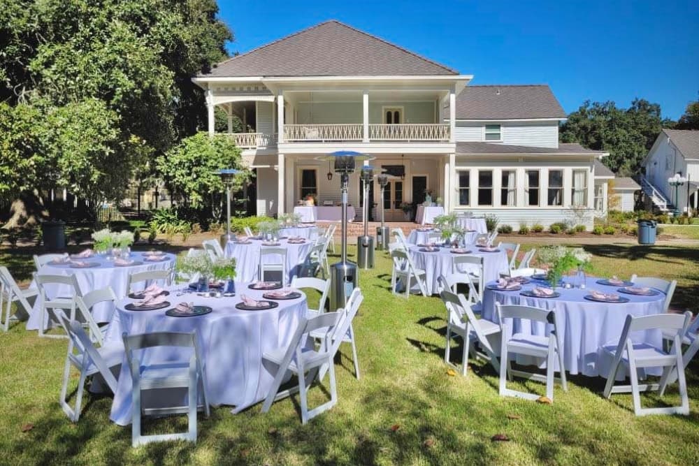 Outdoor dining setup with round tables covered in lavender tablecloths in front of a large, white house.