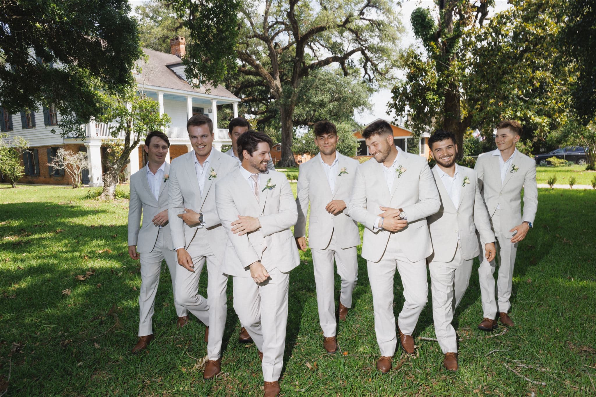 A group of nine men in matching light-colored suits walk together in a lush green yard.