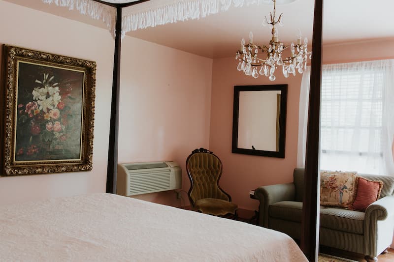 A softly lit pink bedroom featuring a four-poster bed, floral painting, chandelier, and vintage furniture.