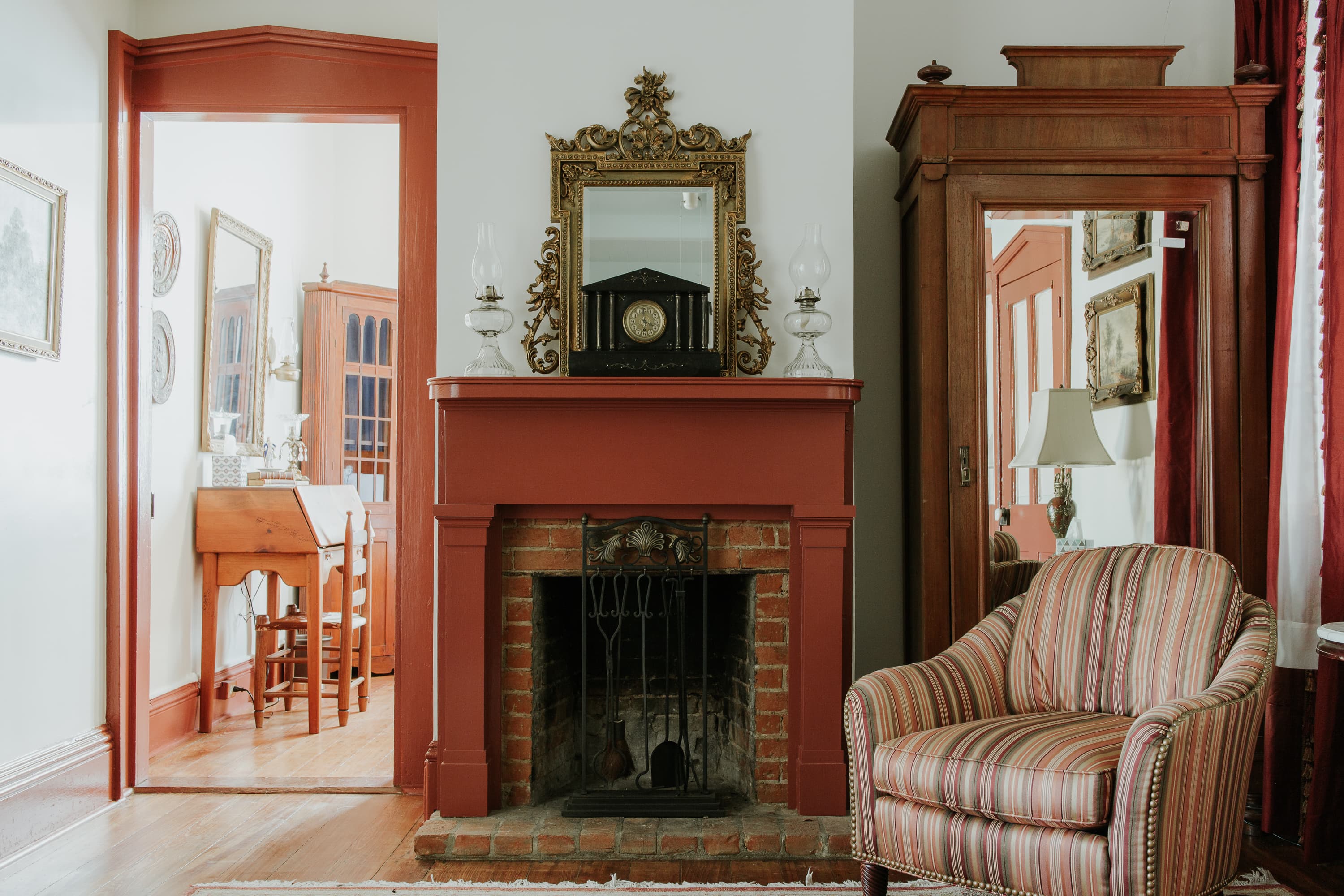 A cozy living room featuring a striped armchair, a red fireplace with a mirror above, and an entrance to a bright adjoining room.