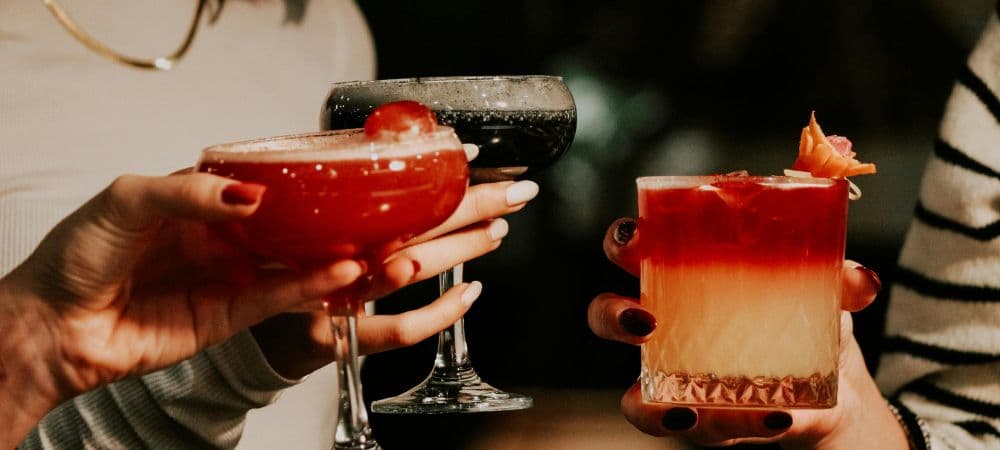 Three hands hold colorful cocktails in a cozy setting.