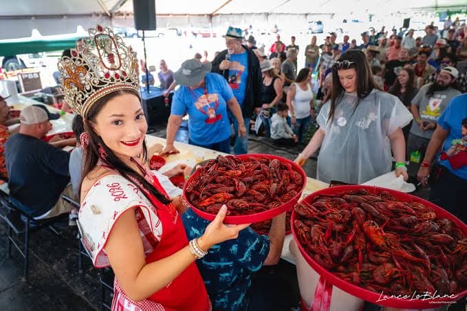 A queen holds two large trays of crawfish in front of a lively crowd at a festival.