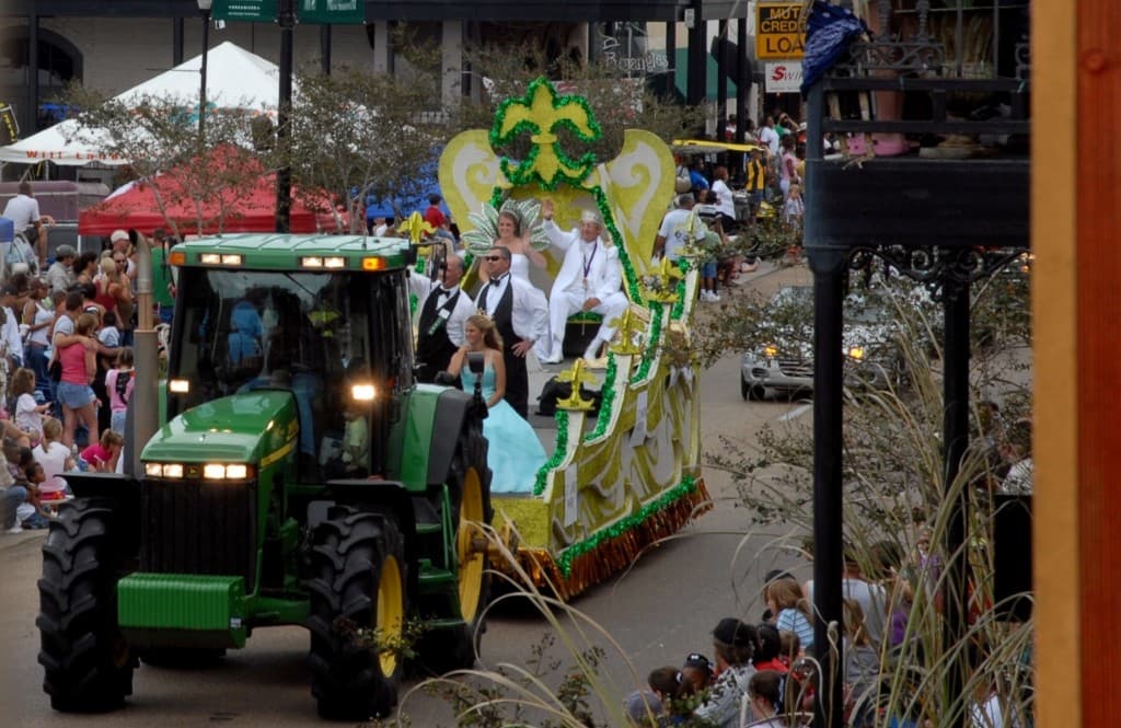 A decorated float pulled by a green tractor moves through a parade with crowds lining the street.