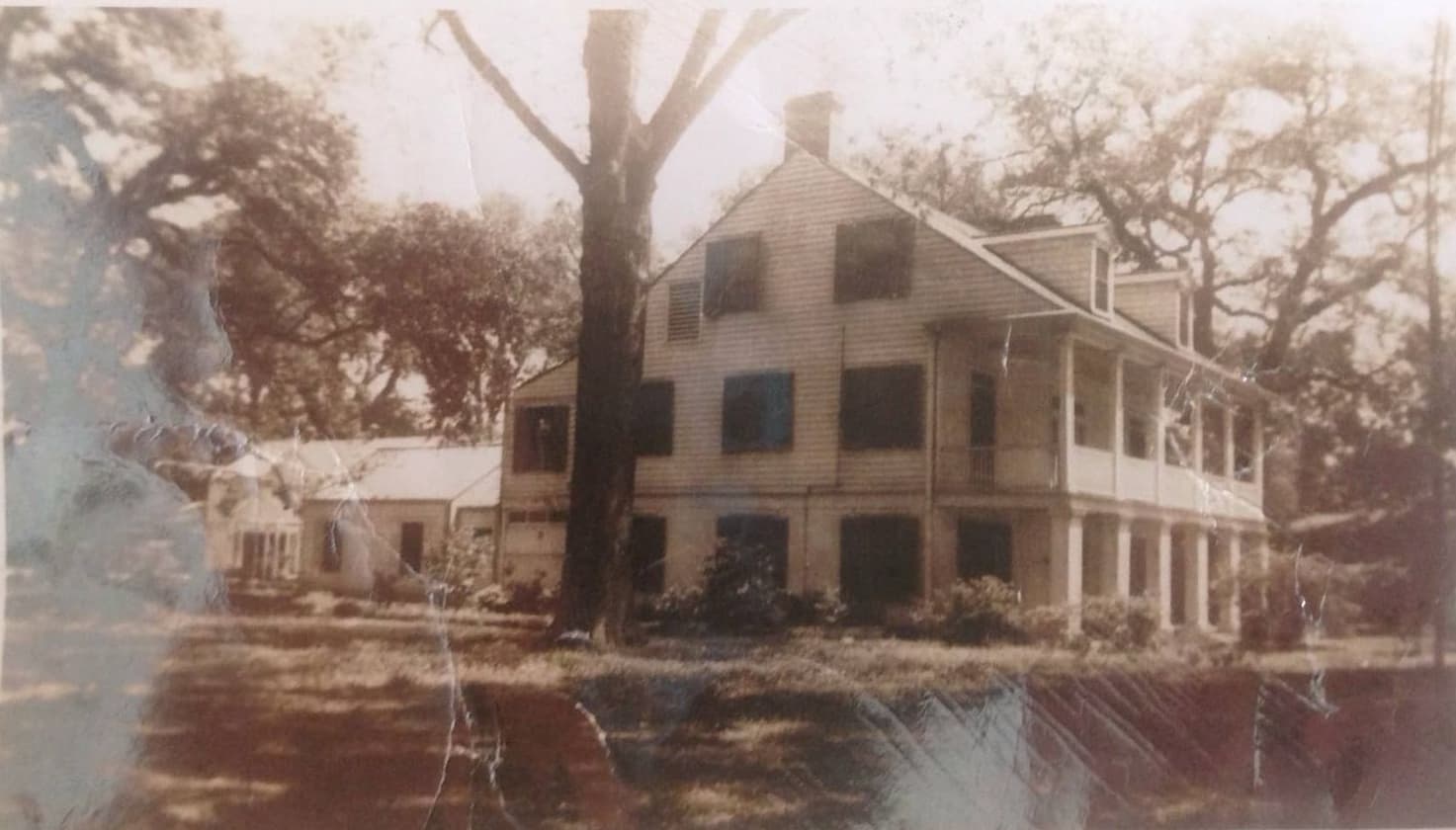 A vintage photograph of a two-story house surrounded by trees and greenery.