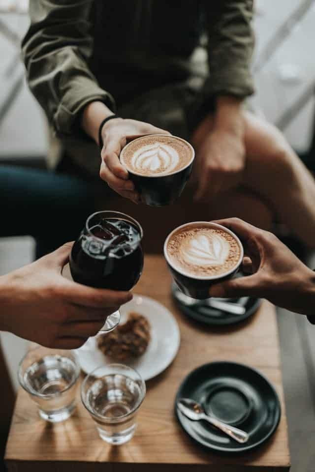 Two hands clink coffee cups above a wooden table with pastries and glasses of water.