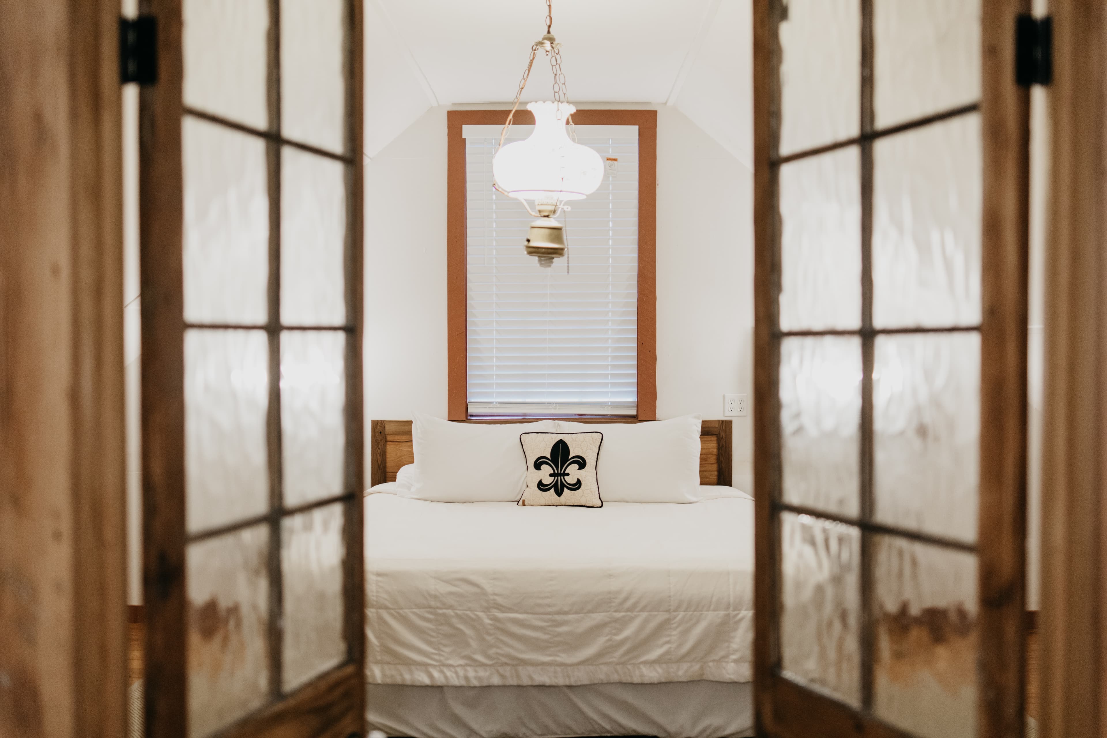 A cozy bedroom viewed through glass-paneled doors, featuring a bed with a decorative pillow and a pendant light overhead.